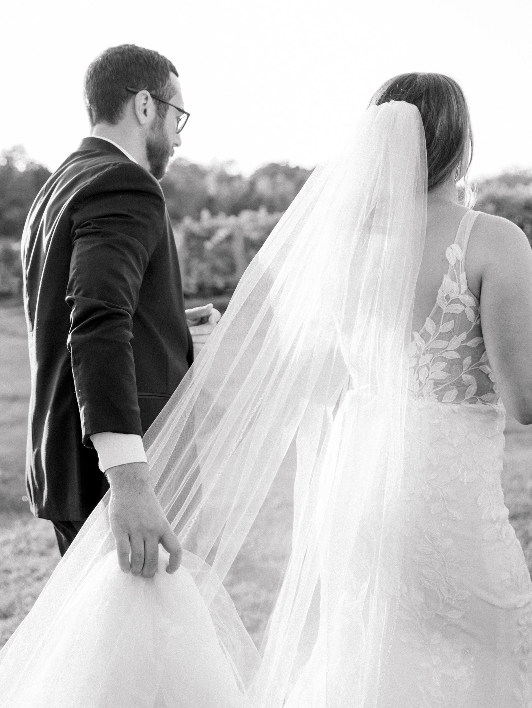 Black and white wedding photo of groom holding the bride’s gown and veil while walking through the vineyard at Ashton Creek Vineyards in Chester VA.