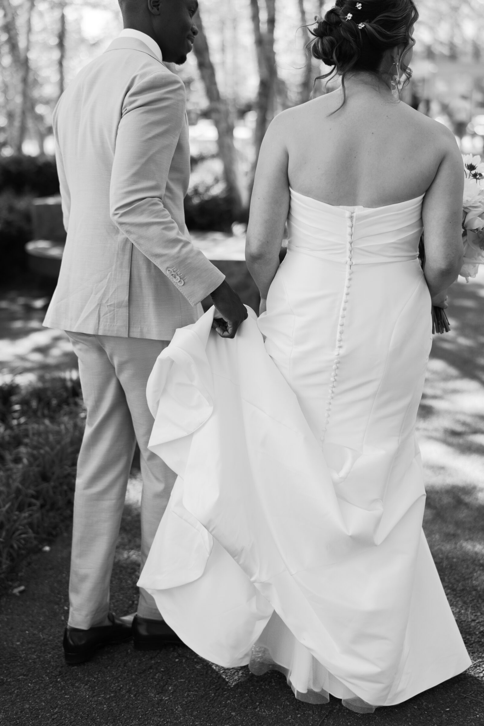 Black and white wedding photo of groom carrying the bride’s dress train as they walk together near District Winery in Washington DC.