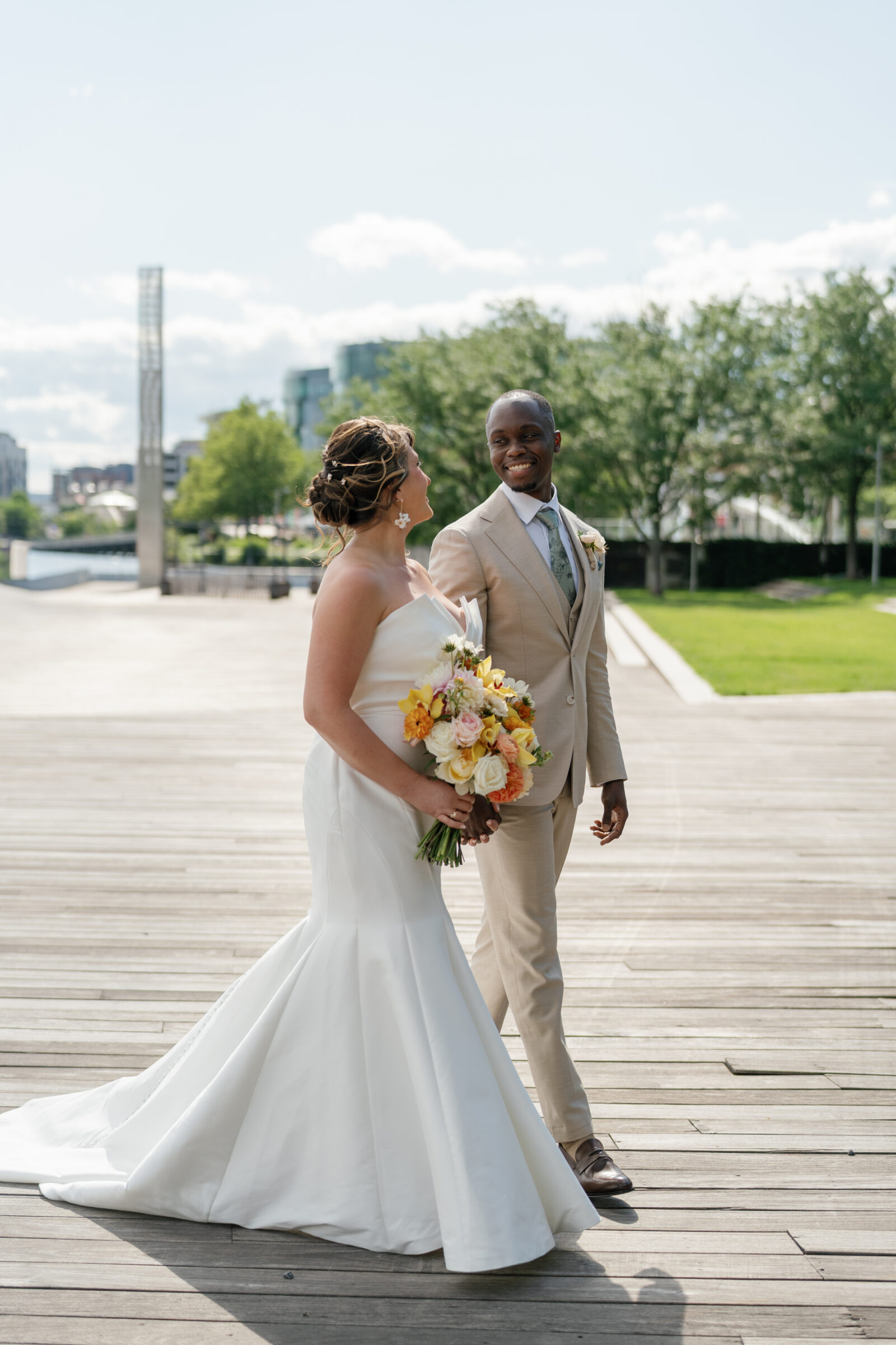 Bride and groom walking hand in hand on the boardwalk near District Winery in Washington DC with waterfront views and modern architecture in the background.