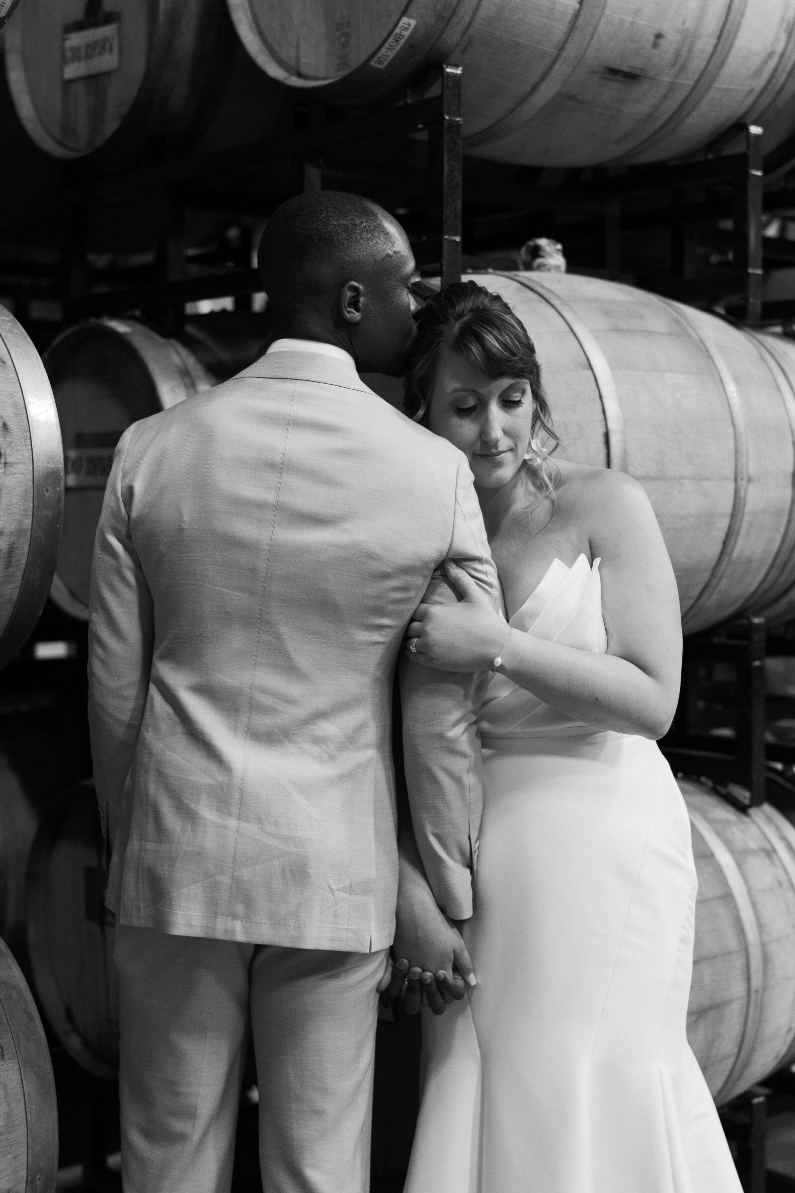 Black and white wedding photo of bride leaning on groom’s shoulder as he kisses her forehead in the barrel room at District Winery in Washington DC.