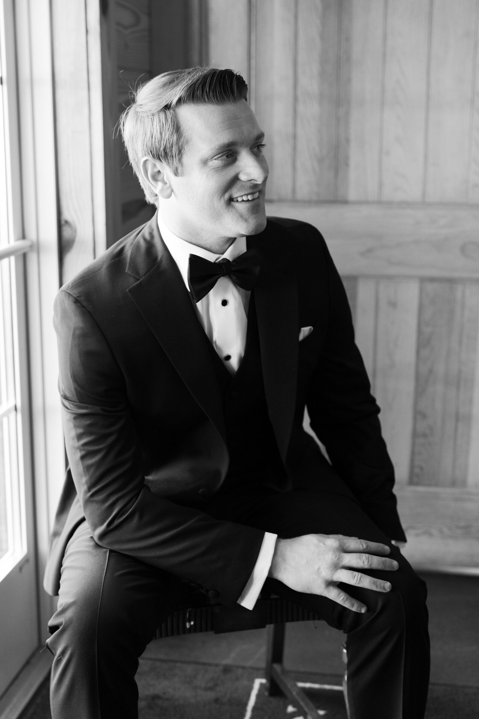 Groom in a black tuxedo and bow tie sitting by a window, smiling as he prepares for a September wedding at Middleburg Barn