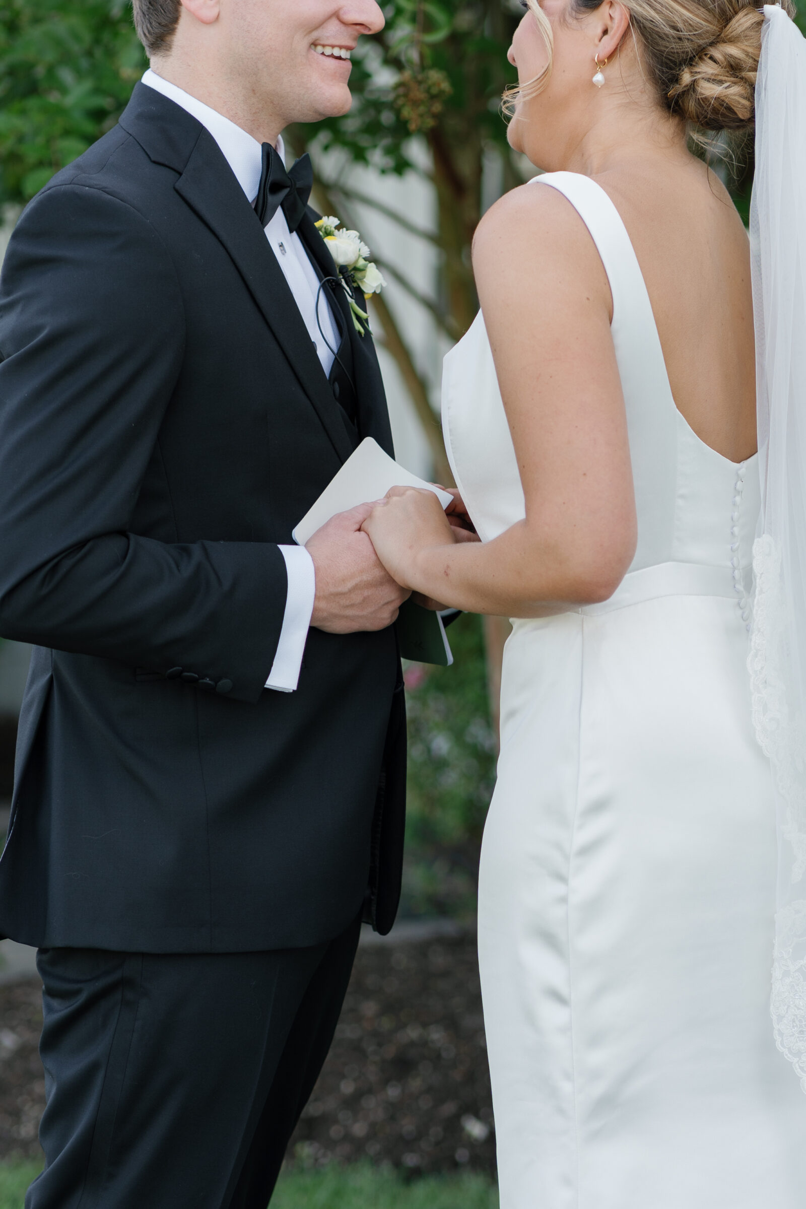 Bride in a simple satin gown and veil and groom in a black tux hold hands with a vow book during their first look at a September wedding at Middleburg Barn