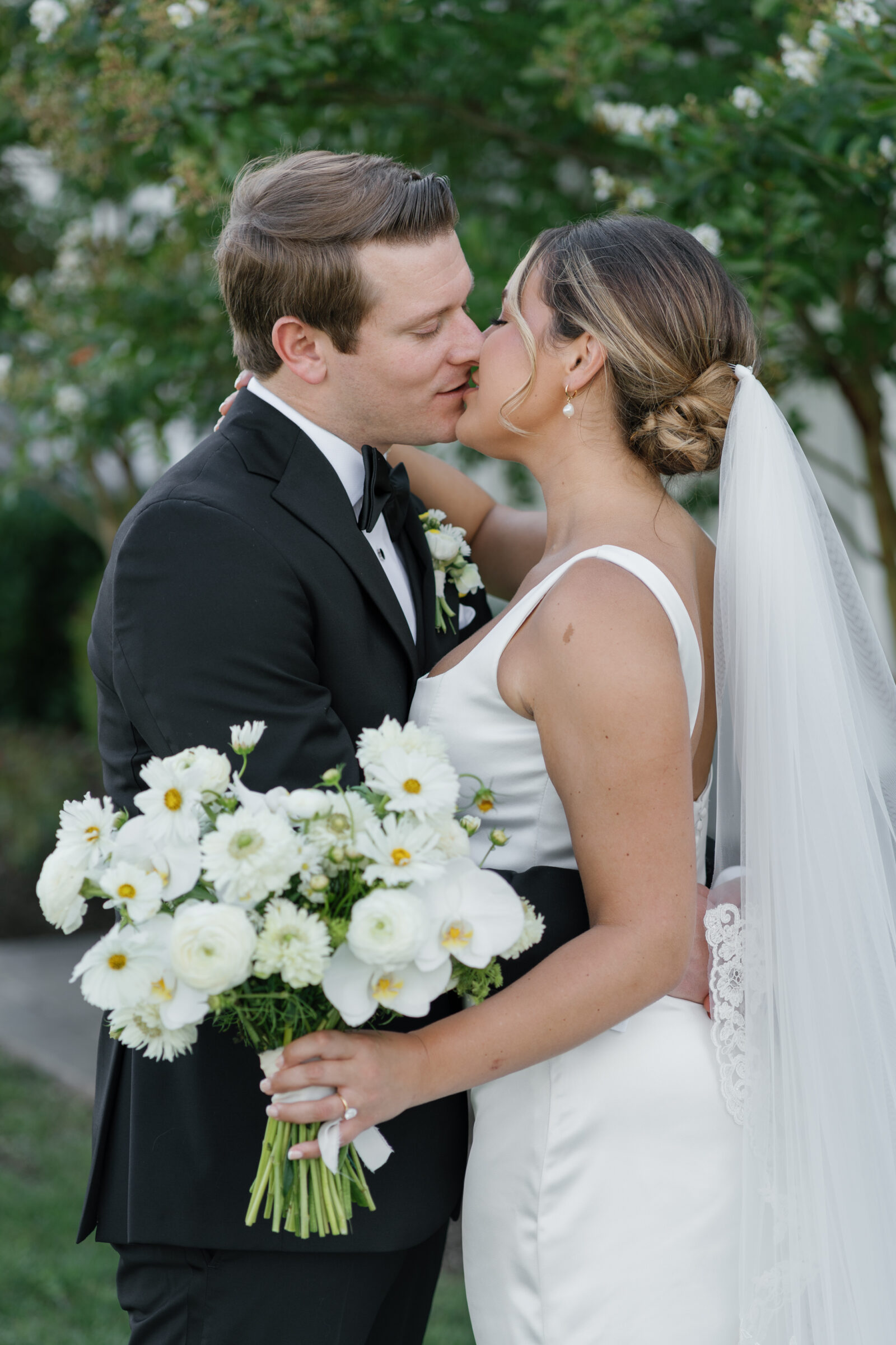 Bride in a simple satin gown and lace veil kisses groom in a black tux as she holds a white garden-style bouquet in the venue’s garden.