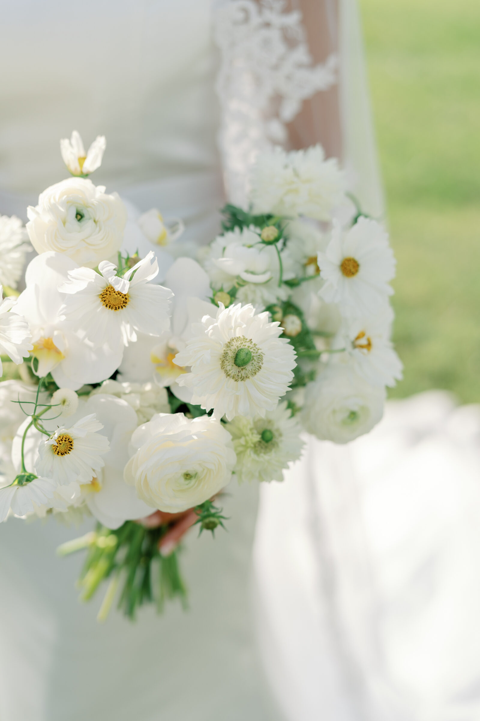 close-up of the bride’s sunlit white garden-style bouquet (ranunculus, anemones, cosmos and orchids) held against her satin gown and lace veil on Middleburg Barn’s lawn