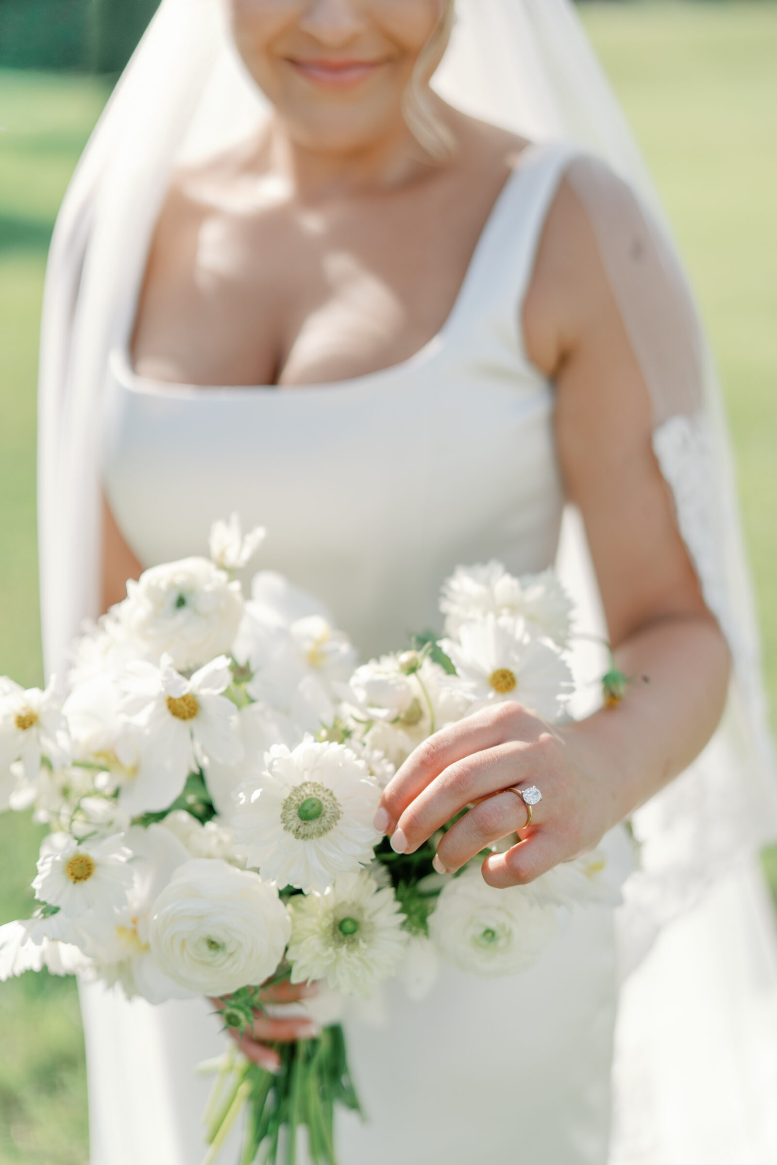 close-up of the bride in a square-neck satin gown and lace veil holding a sunlit white garden bouquet (ranunculus, anemones, cosmos) while showing her engagement ring