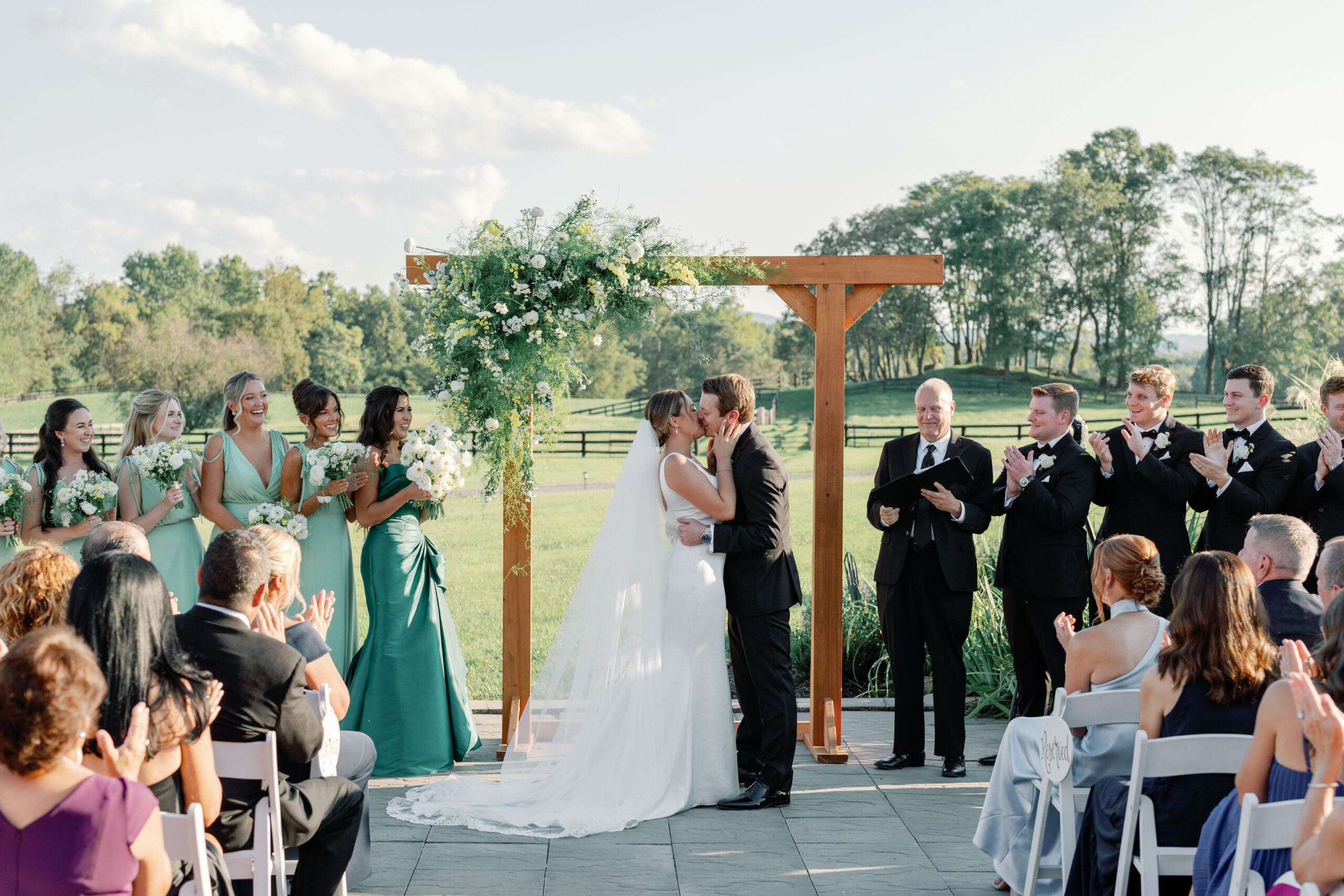 Middleburg Barn — bride and groom kiss under a wooden floral arch as bridesmaids in green and guests applaud on a sunlit Virginia lawn.