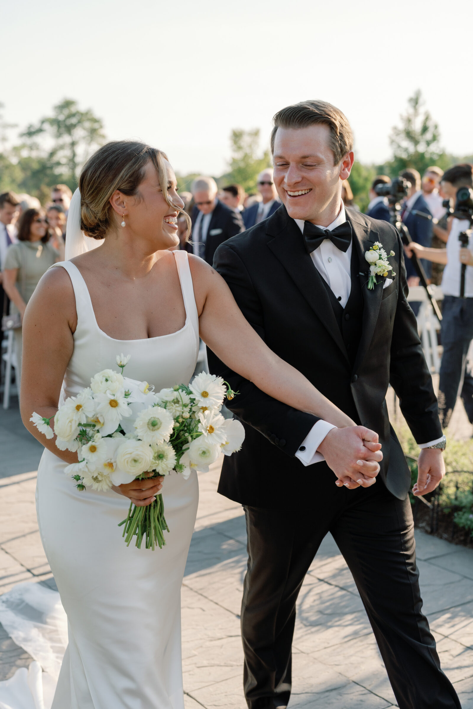 Wedding at Middleburg Barn in Virginia — bride in a satin gown holding a white ranunculus bouquet and groom in a black tux walk hand-in-hand down the aisle, smiling as guests applaud.