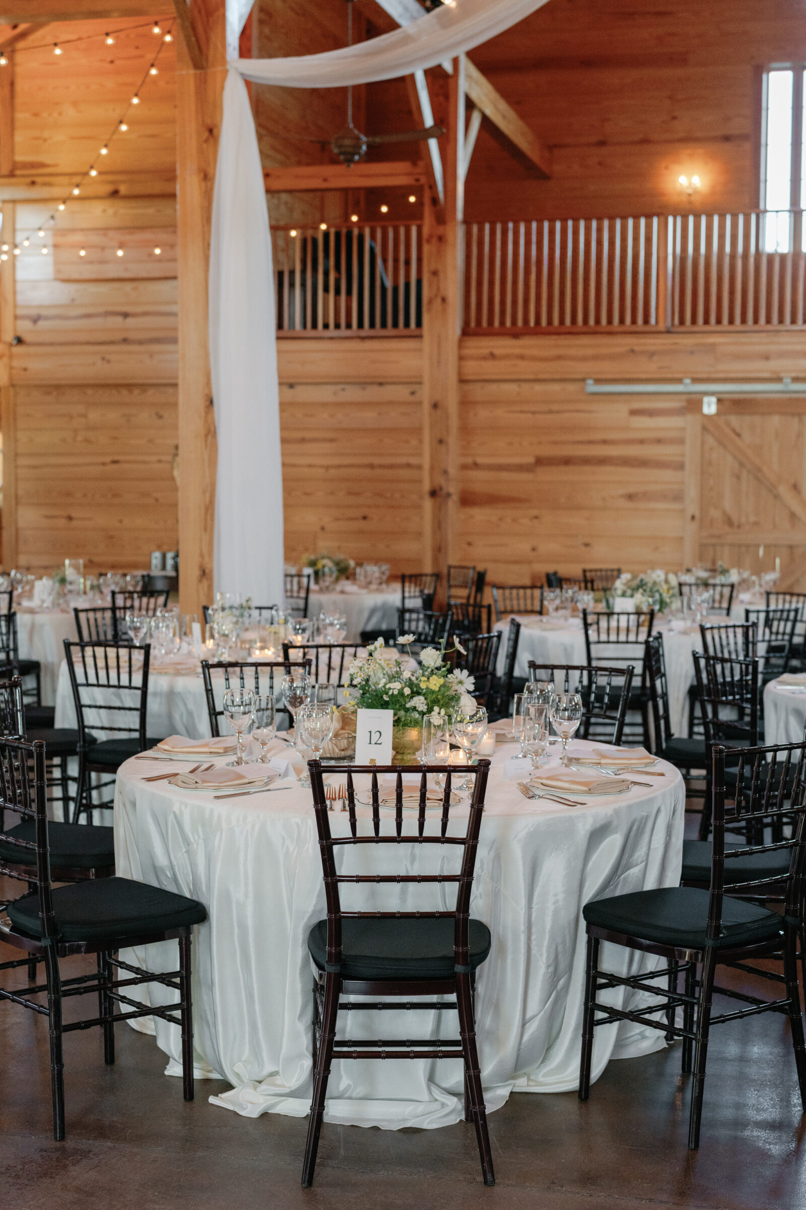 Reception tables with white linens and dark chiavari chairs set for a September wedding at Middleburg Barn, wooden beams, string lights, and floral centerpieces around table number 12.