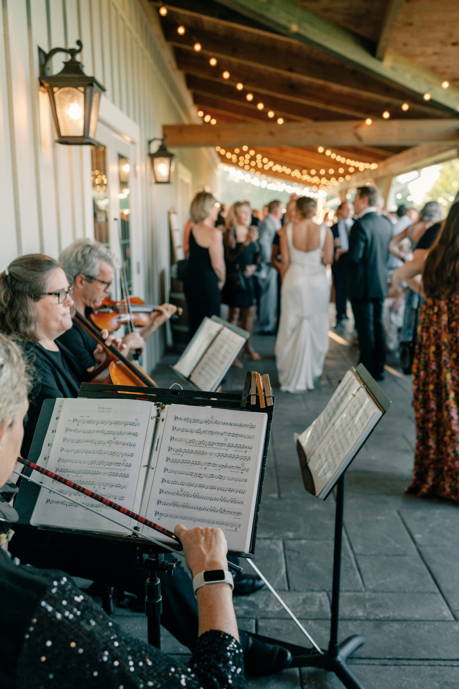 String quartet playing from music stands on a covered porch while the bride and groom mingle with guests under bistro lights.