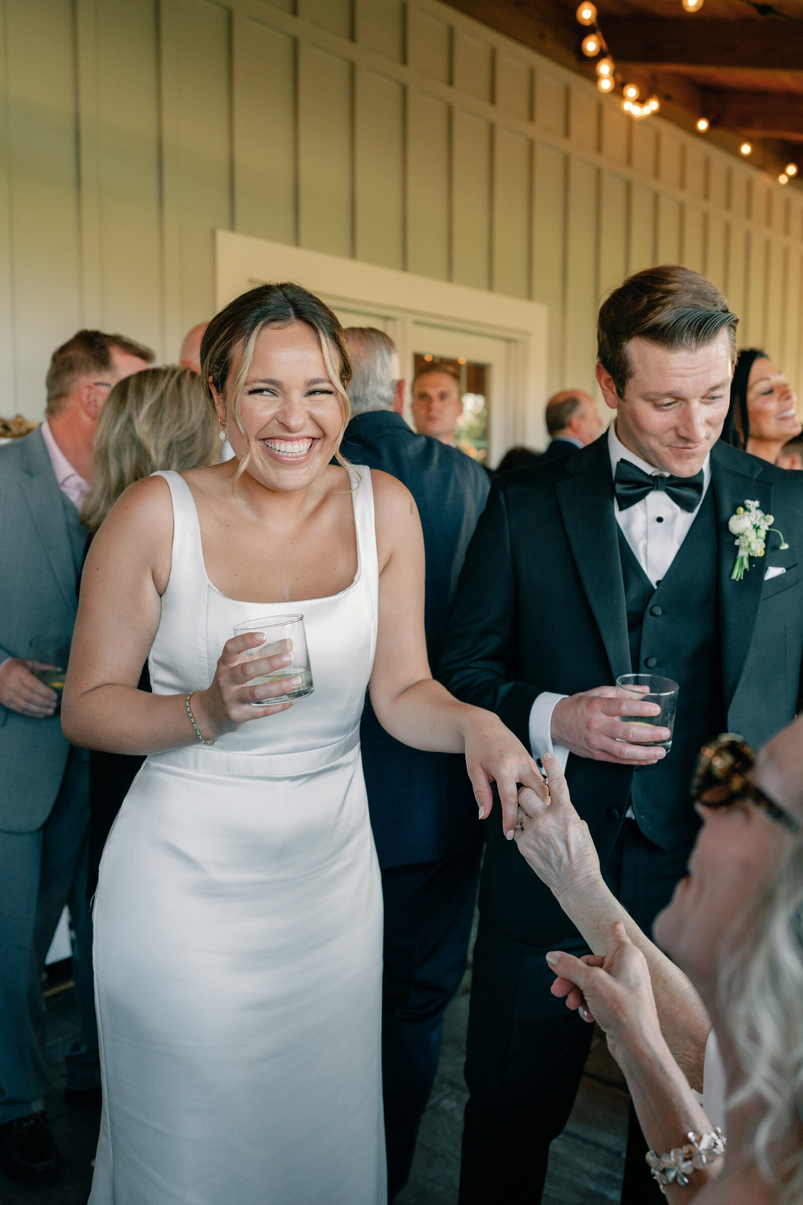 Bride in a silk gown laughs with her groom in a black tux while holding a guest’s hand and drinks beneath bistro lights on the covered porch during the reception — wedding at Middleburg Barn.