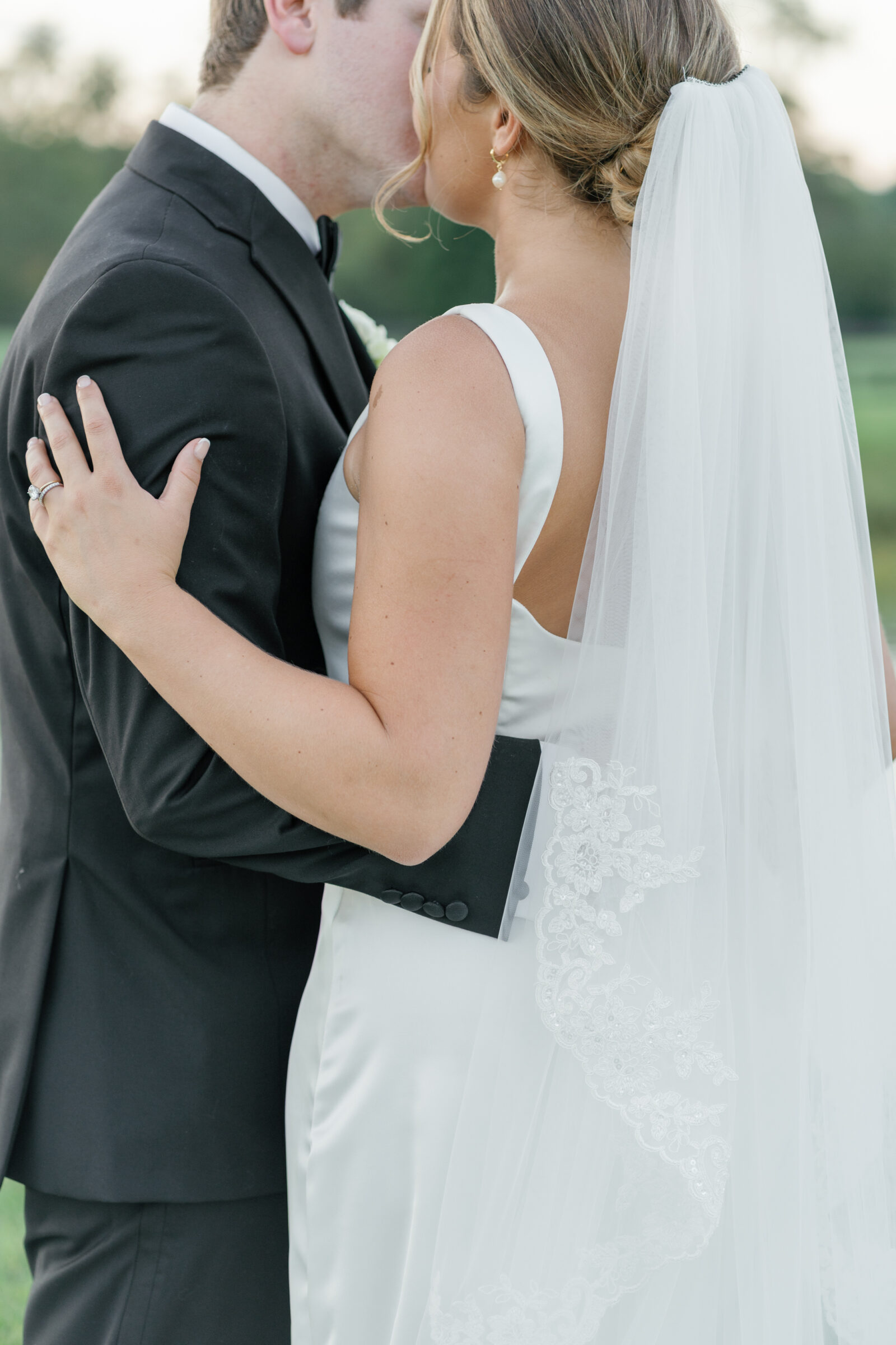 Bride and groom share a gentle kiss as they embrace in a field — bride in a satin gown with a lace-edged veil and groom in a classic black tux, a romantic moment from a wedding at Middleburg Barn.