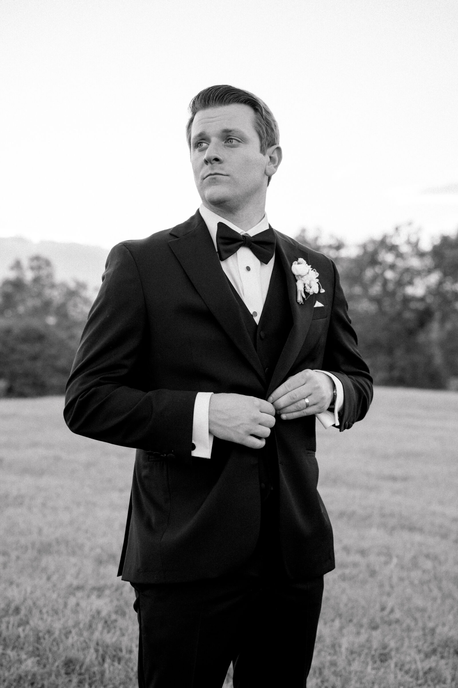 Groom in a black tux and bow tie buttoning his jacket in a field — classic portrait from a wedding at Middleburg Barn.