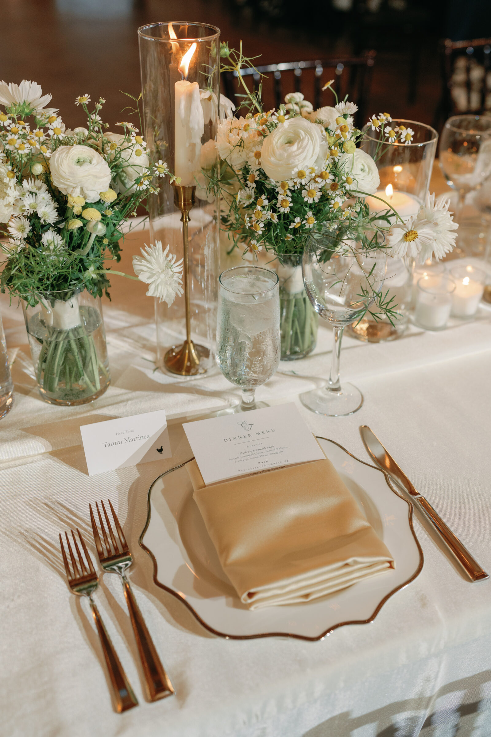 Elegant reception place setting at a September Wedding at Middleburg Barn — scalloped white china with a gold rim, folded gold satin napkin topped with a printed menu card, water goblet and gold flatware, surrounded by small white ranunculus and chamomile floral arrangements, votive candles and a tall glass hurricane candle on a white linen table.