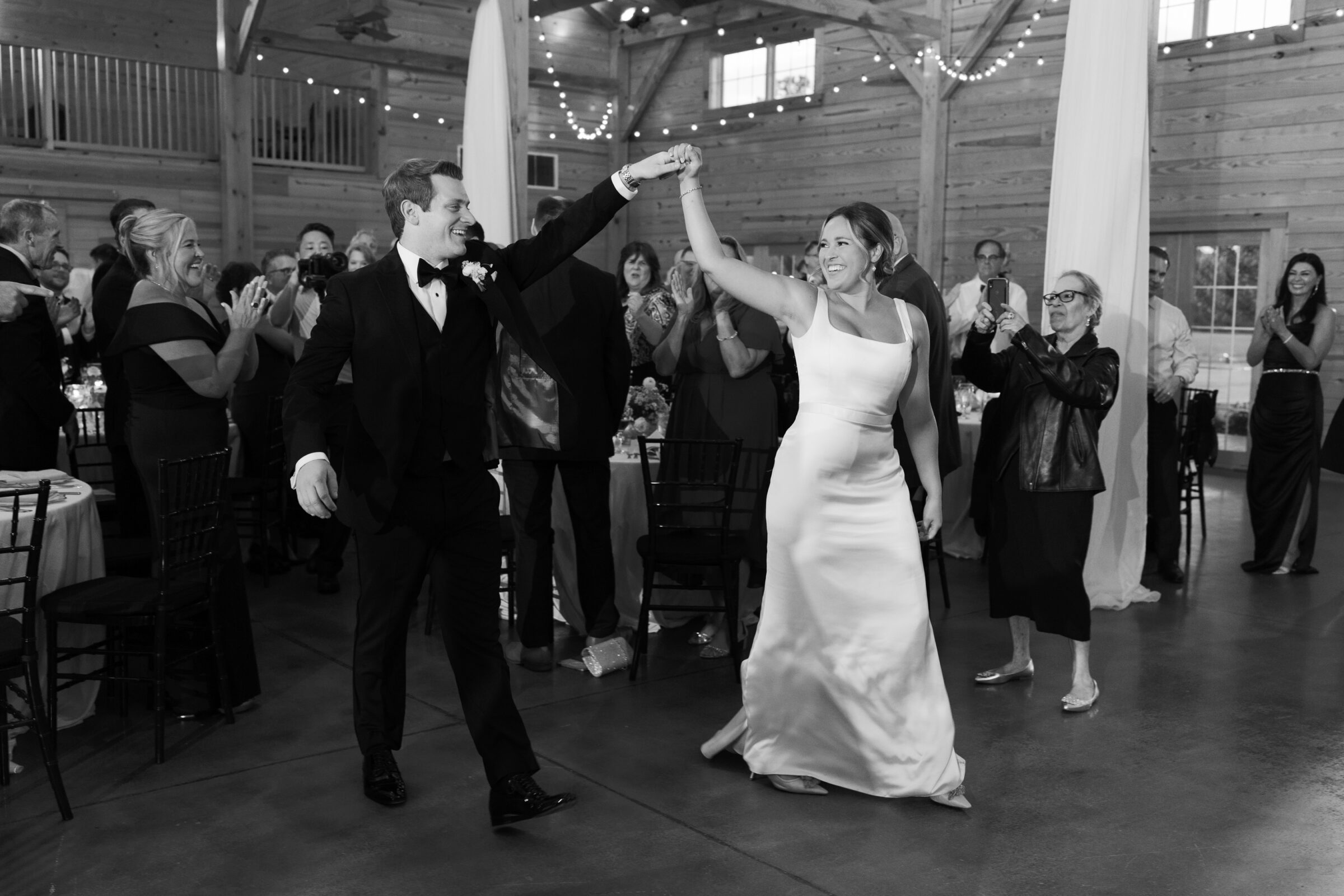 Bride and groom twirling into their reception under string lights in the rustic Middleburg Barn, bride in a satin gown and veil and groom in a black tuxedo as guests cheer — Wedding at Middleburg Barn.