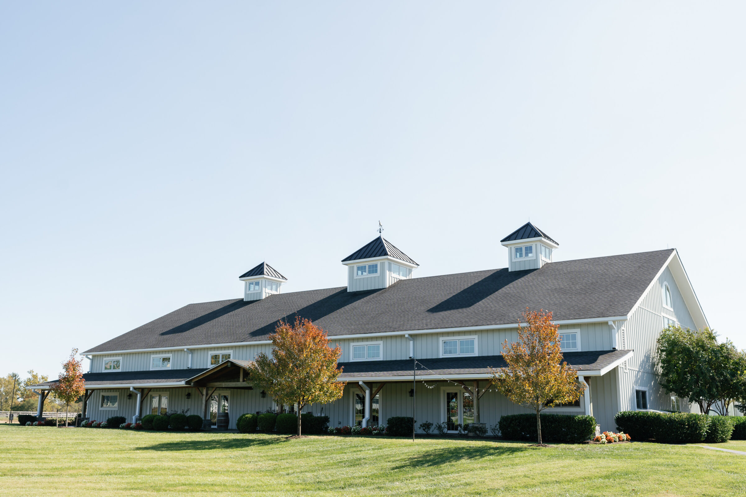 Wide exterior of the Middleburg Barn wedding venue: a long white timber barn with three cupolas, covered porch, string lights and autumn trees set on a manicured green lawn under a clear blue sky — perfect setting for a Wedding at Middleburg Barn.