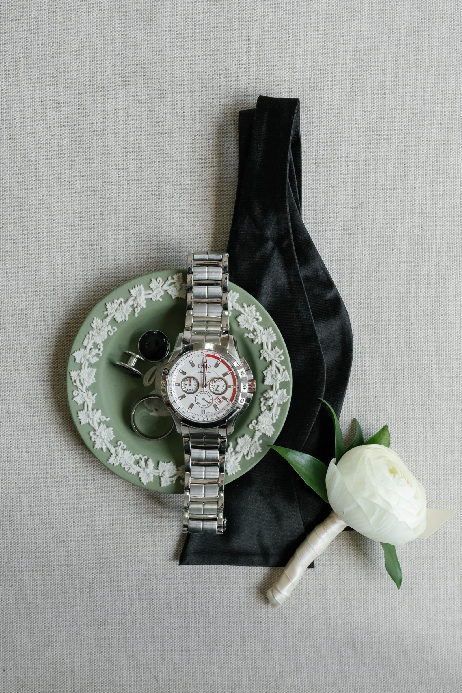 Groom’s wedding details including a silver wristwatch, black cufflinks, wedding band, black bow tie, and white boutonniere styled on a sage Wedgwood plate