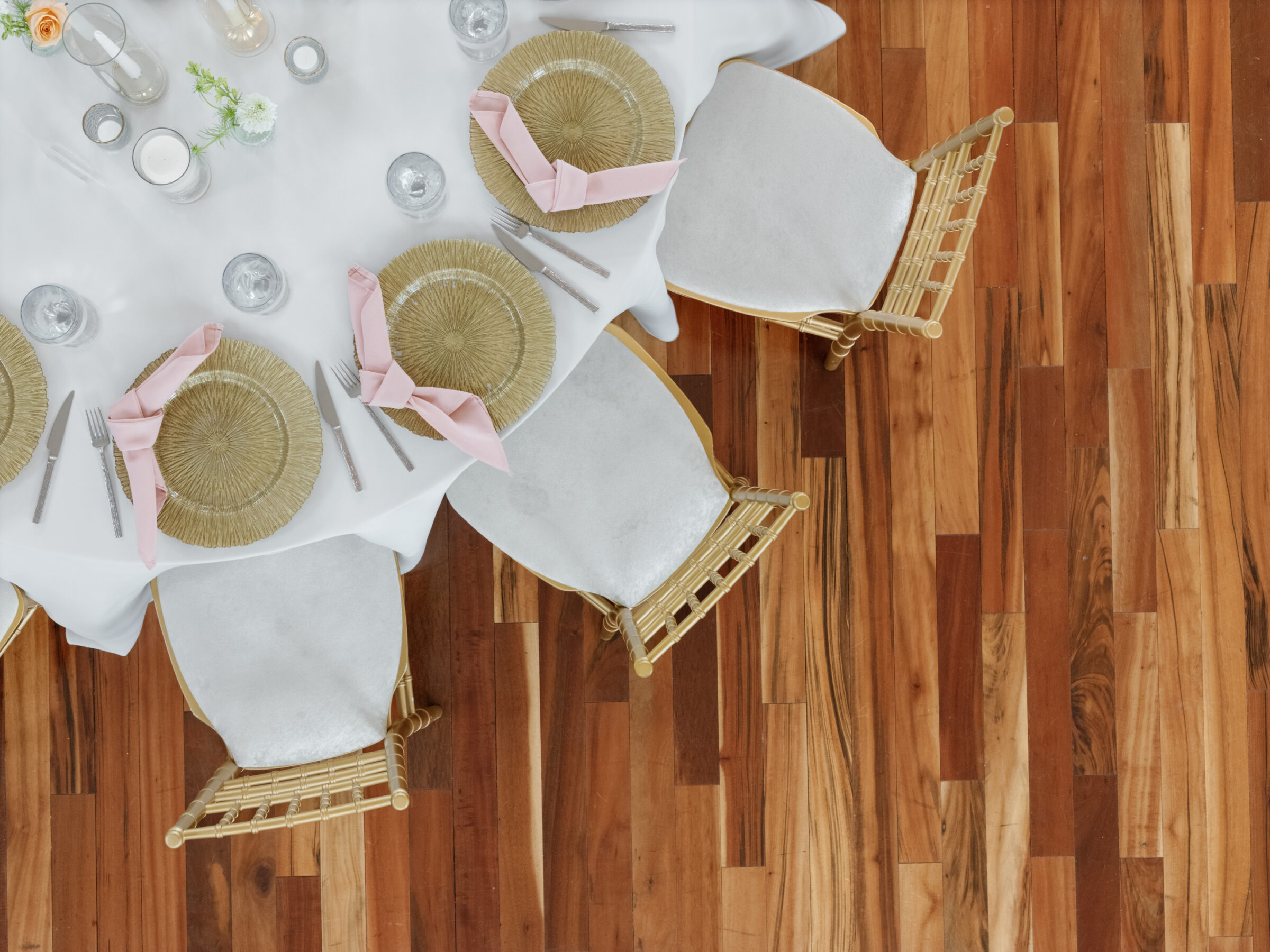 Overhead view of a wedding reception table at The View at Bluemont, a Northern Virginia wedding venue, featuring gold chargers with blush pink napkins, elegant glassware, and gold chiavari chairs on warm wood flooring.