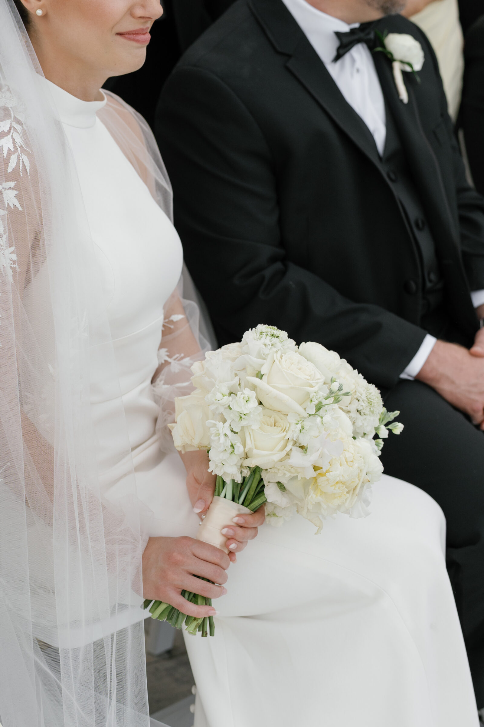 Bride holding a classic white wedding bouquet of roses and peonies while seated with her groom