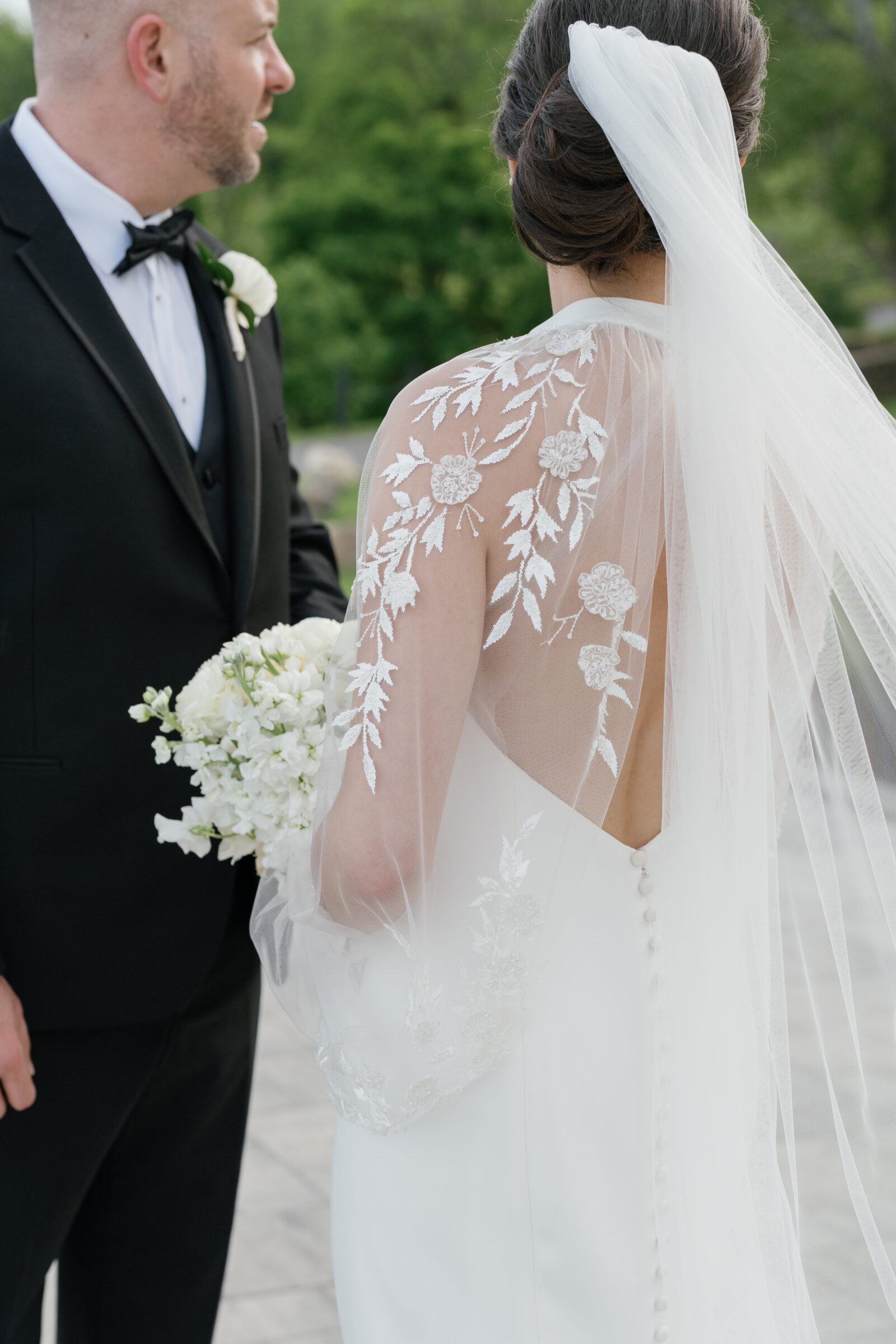 Bride in an elegant crepe gown with illusion lace sleeves and button-back detail standing beside her groom