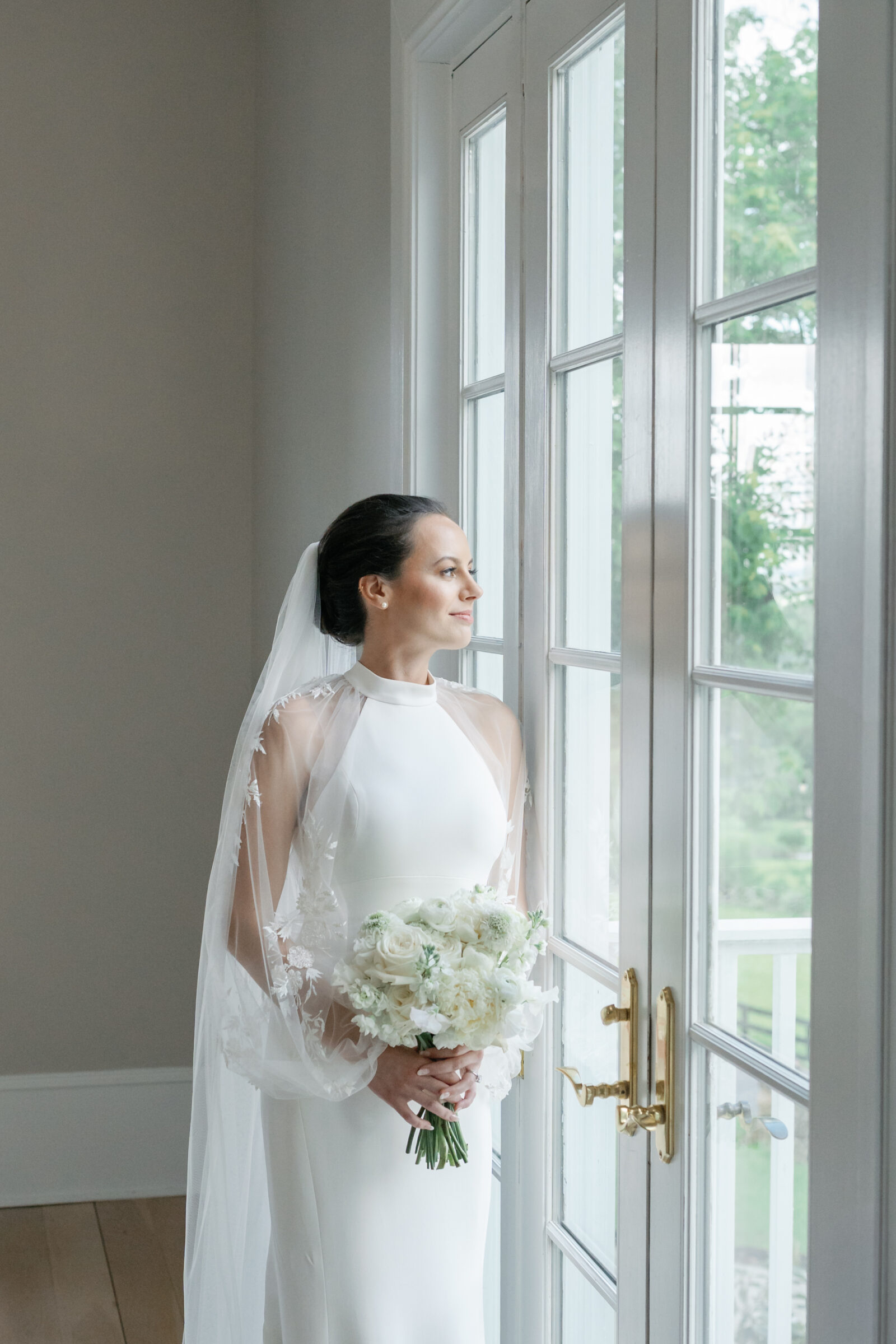 Bride holding a white bouquet, gazing out the window in the bridal suite at The View at Bluemont, an elegant Northern Virginia wedding venue.