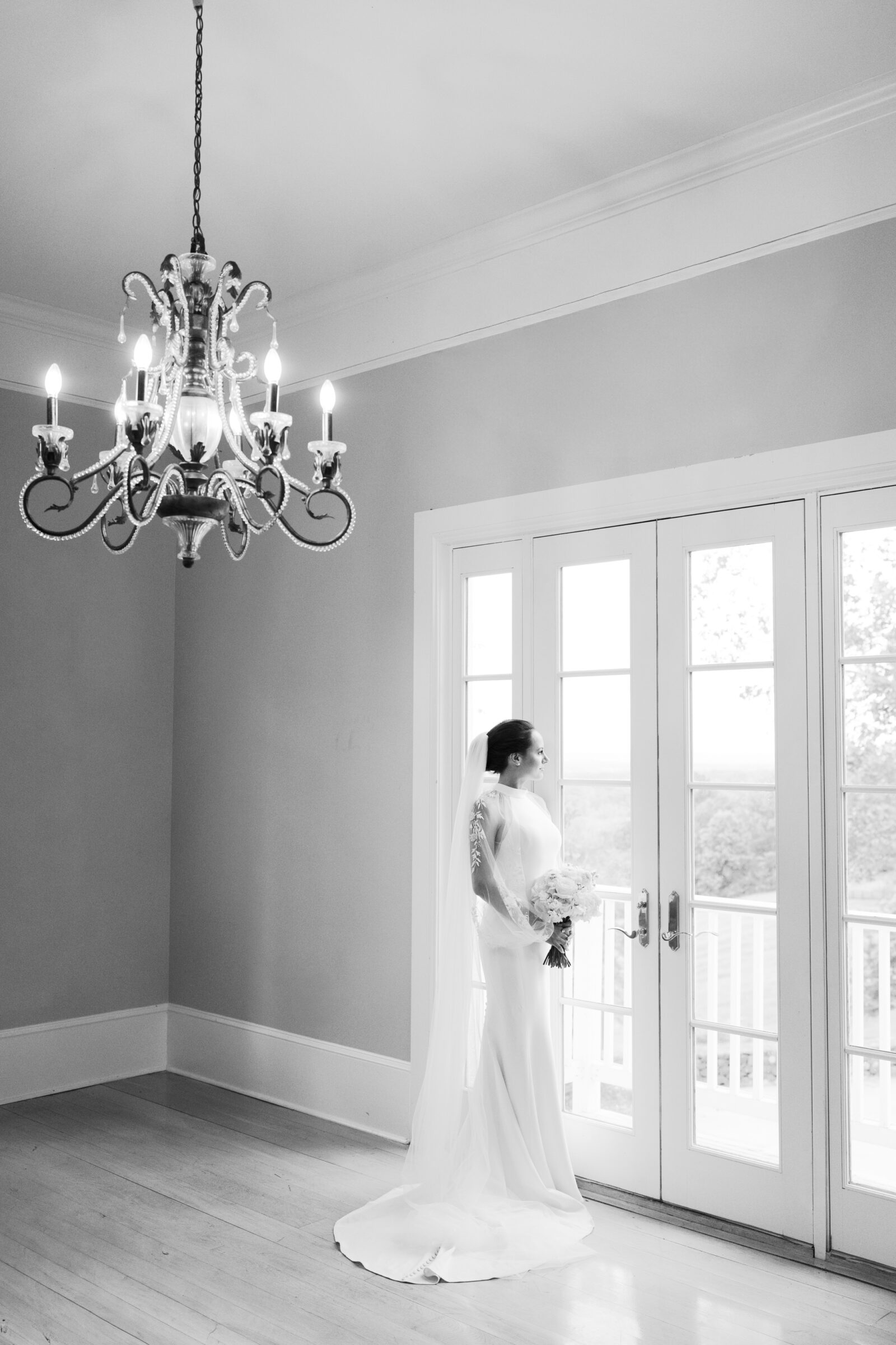 Black and white bridal portrait of a bride holding her bouquet and gazing out French doors beneath an elegant chandelier at The View at Bluemont, a Northern Virginia wedding venue.