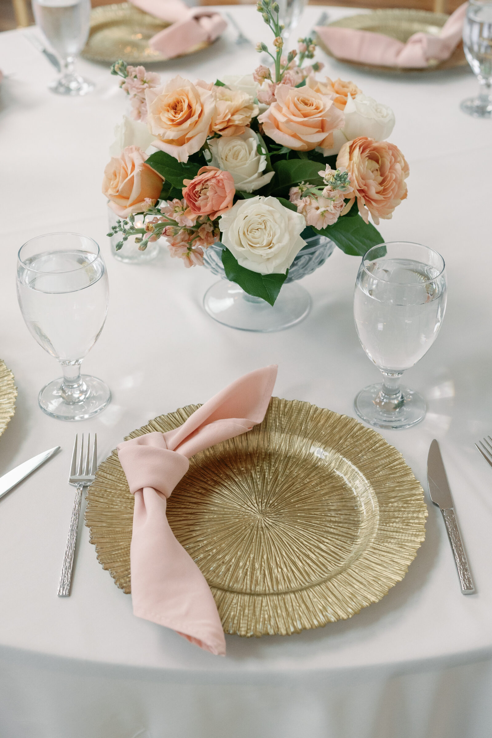 Elegant wedding reception table, featuring a gold charger plate with a blush pink napkin and a centerpiece of peach and white roses.