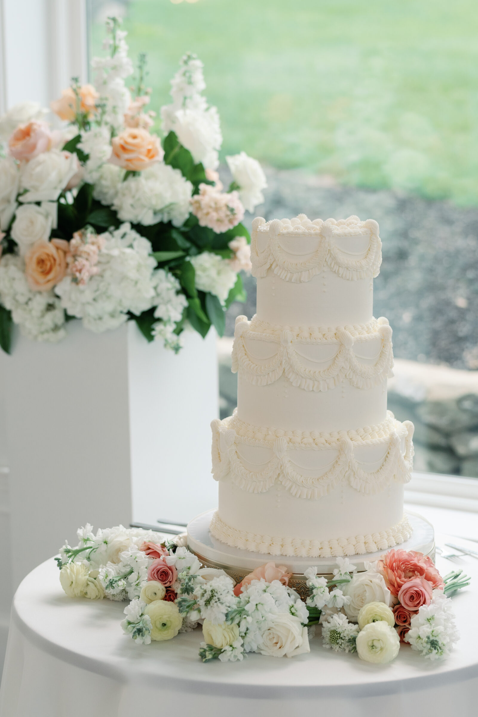 Elegant three-tier white wedding cake with piped vintage-inspired detailing, surrounded by blush, peach, and white floral arrangements.