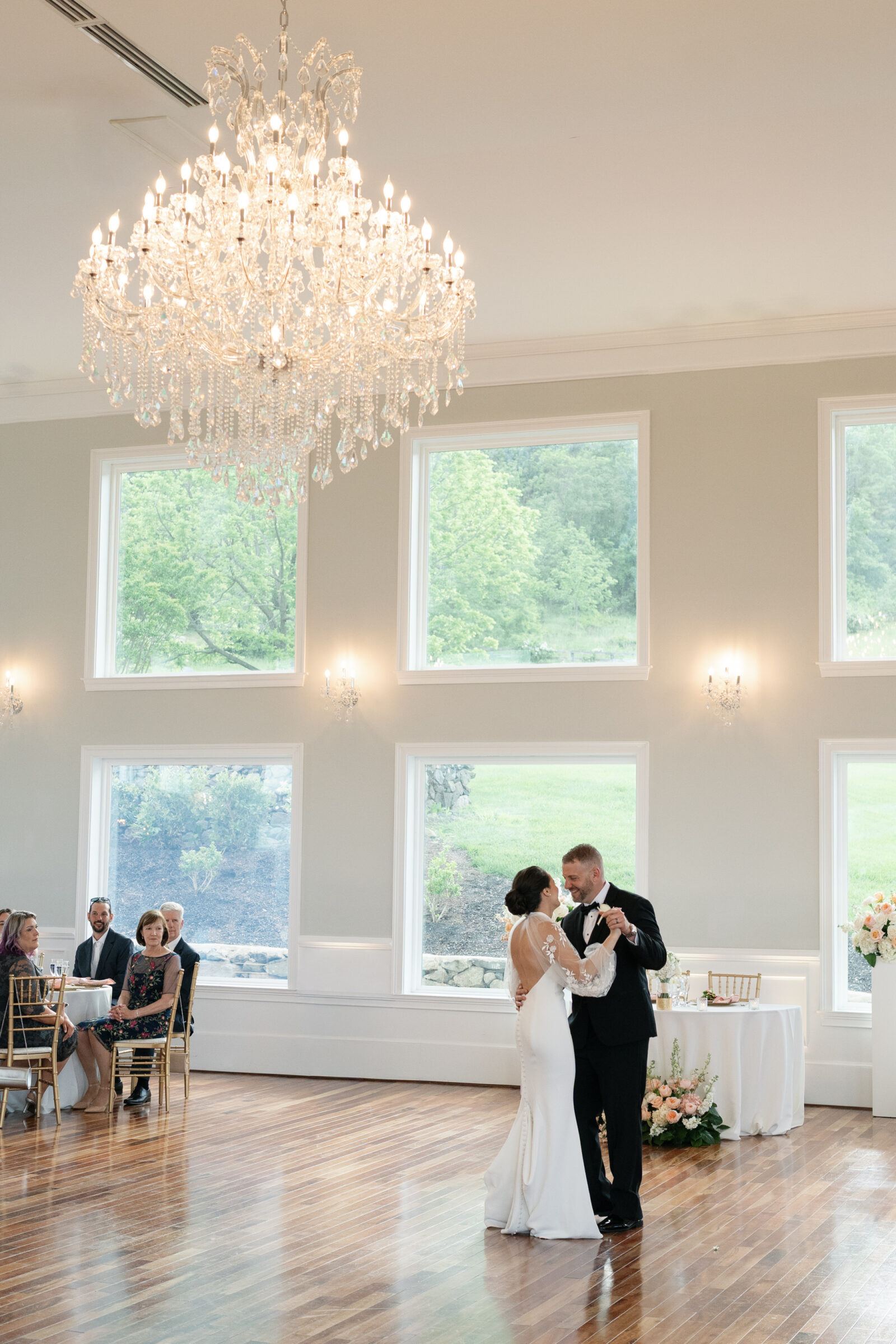 Bride and groom share their first dance beneath a sparkling chandelier in the grand ballroom at The View at Bluemont, a Northern Virginia wedding venue, surrounded by guests and floor-to-ceiling windows overlooking the countryside.