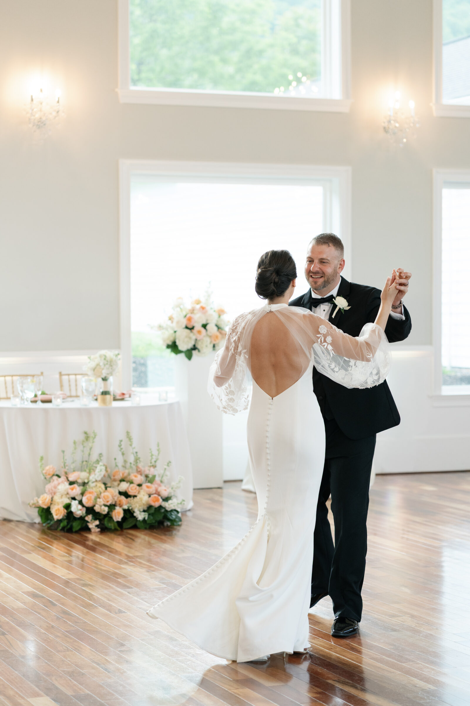 Bride and groom share a joyful first dance in the ballroom at The View at Bluemont, a Northern Virginia wedding venue, with romantic floral arrangements and floor-to-ceiling windows framing the scene.