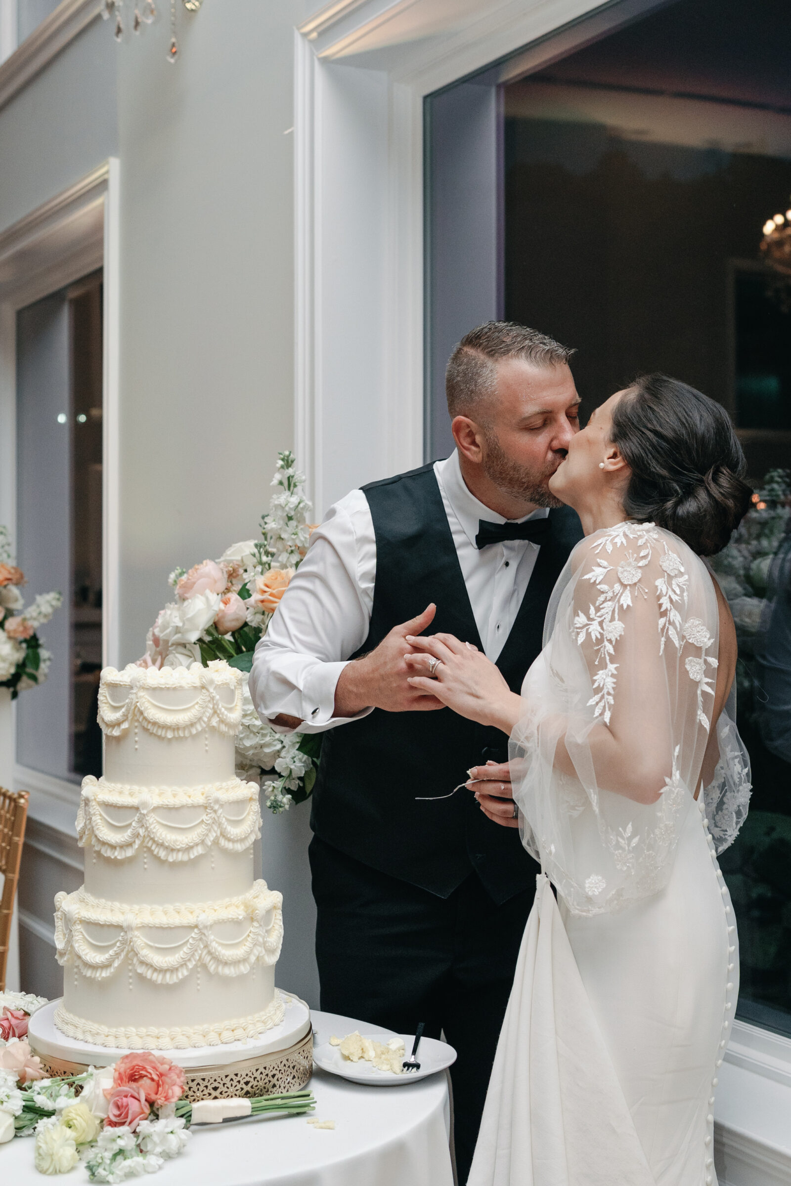 Bride and groom share a kiss after cutting their elegant three-tier wedding cake, surrounded by pastel florals at The View at Bluemont, a romantic Northern Virginia wedding venue.