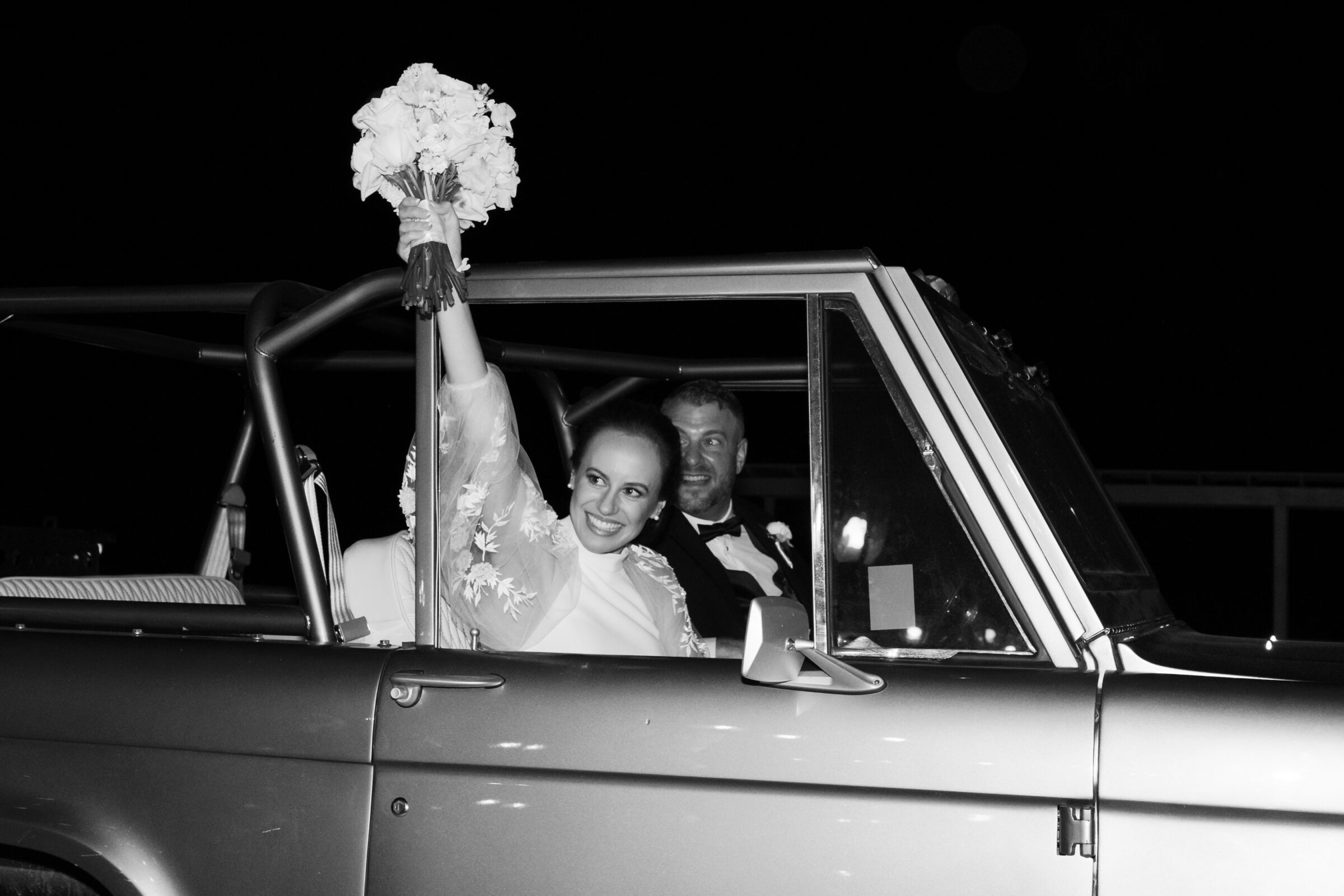 Bride and groom leaving The View at Bluemont in a vintage getaway car, with the bride raising her bouquet in celebration.