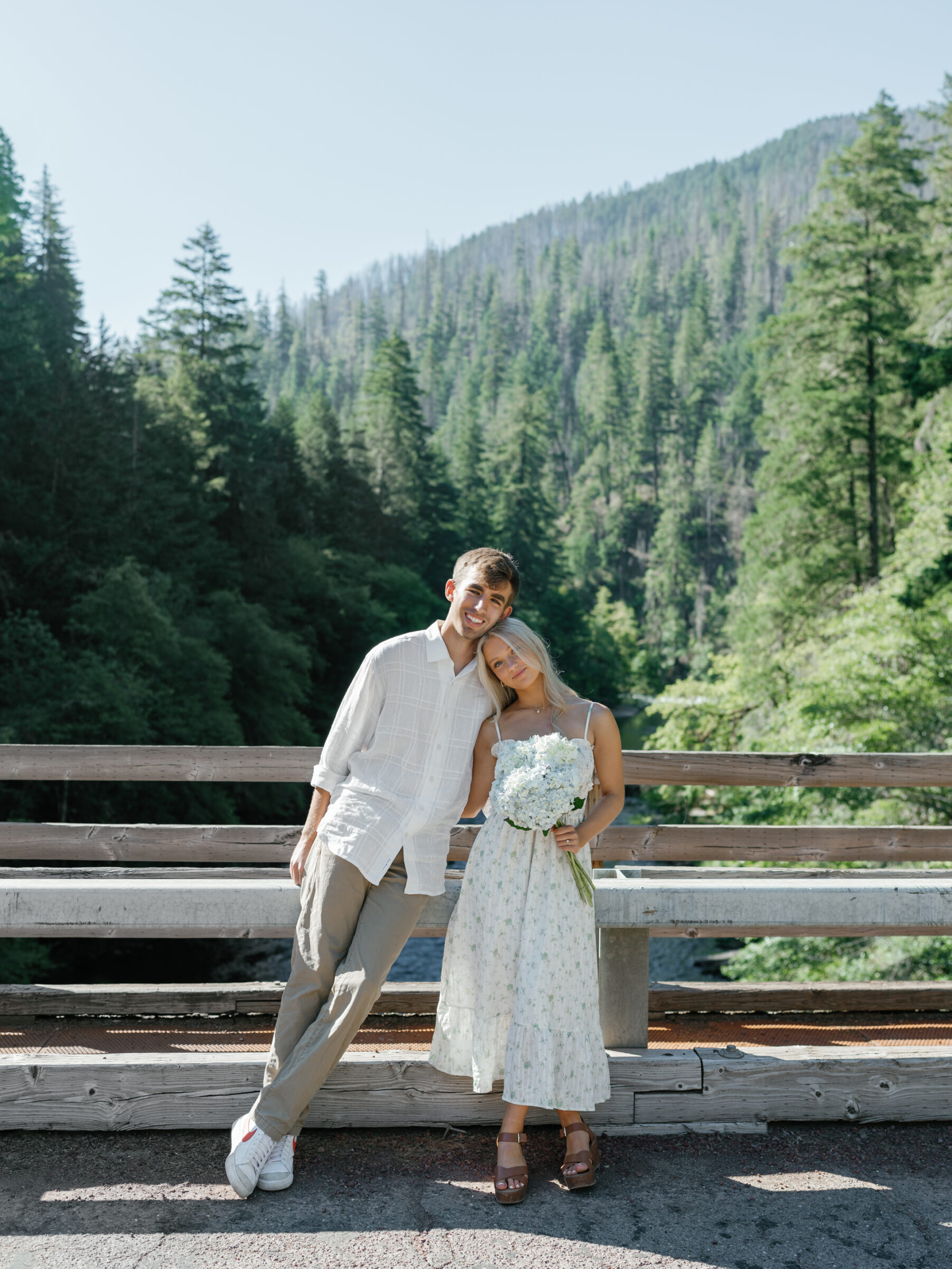 Couple standing on a wooden bridge during their Umpqua River engagement session, surrounded by lush Oregon forest, holding hydrangeas in soft morning light.