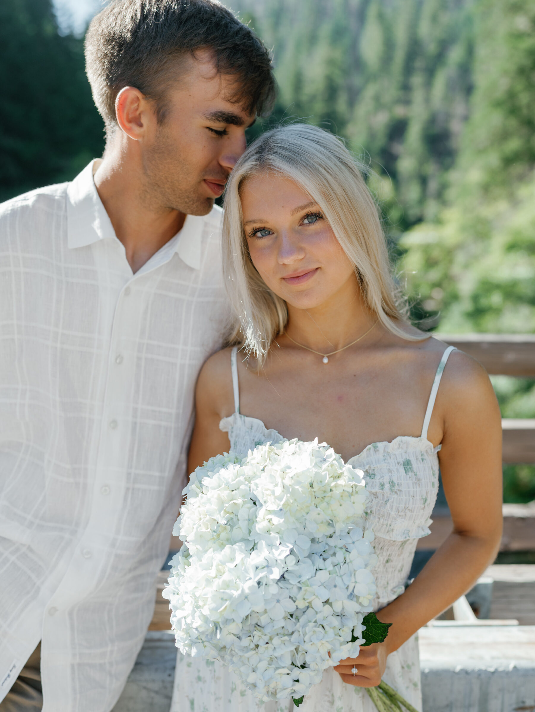 Close-up of engaged couple during their Umpqua River engagement, with the bride-to-be holding a bouquet of white hydrangeas and forest in the background.