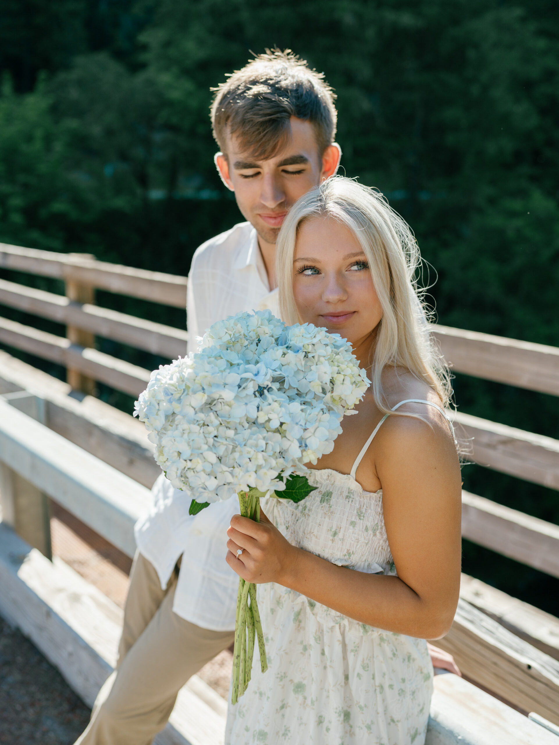 Bride-to-be holding white hydrangeas during an Umpqua River engagement session, with her fiancé standing behind her on a sunlit wooden bridge in Oregon.
