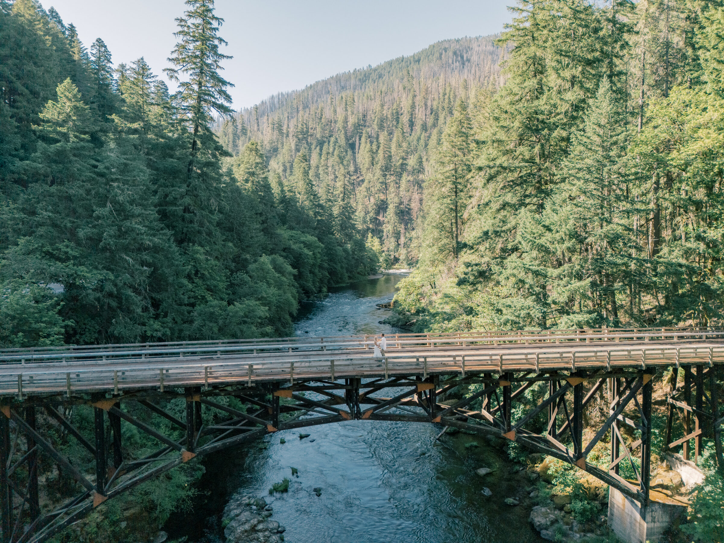 Aerial view of an Umpqua River engagement session in Oregon, showing the couple standing on a rustic wooden bridge surrounded by lush evergreen forest.