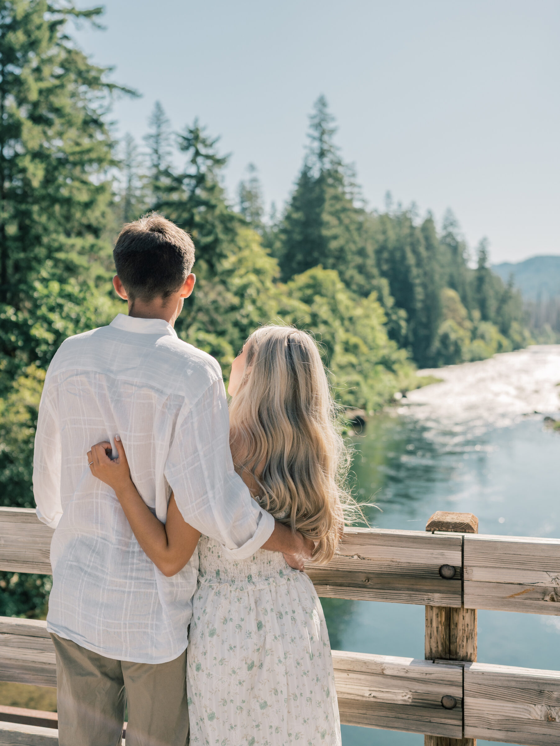 Engaged couple embracing while looking out over the Umpqua River during their Oregon engagement session, framed by evergreens and flowing water.