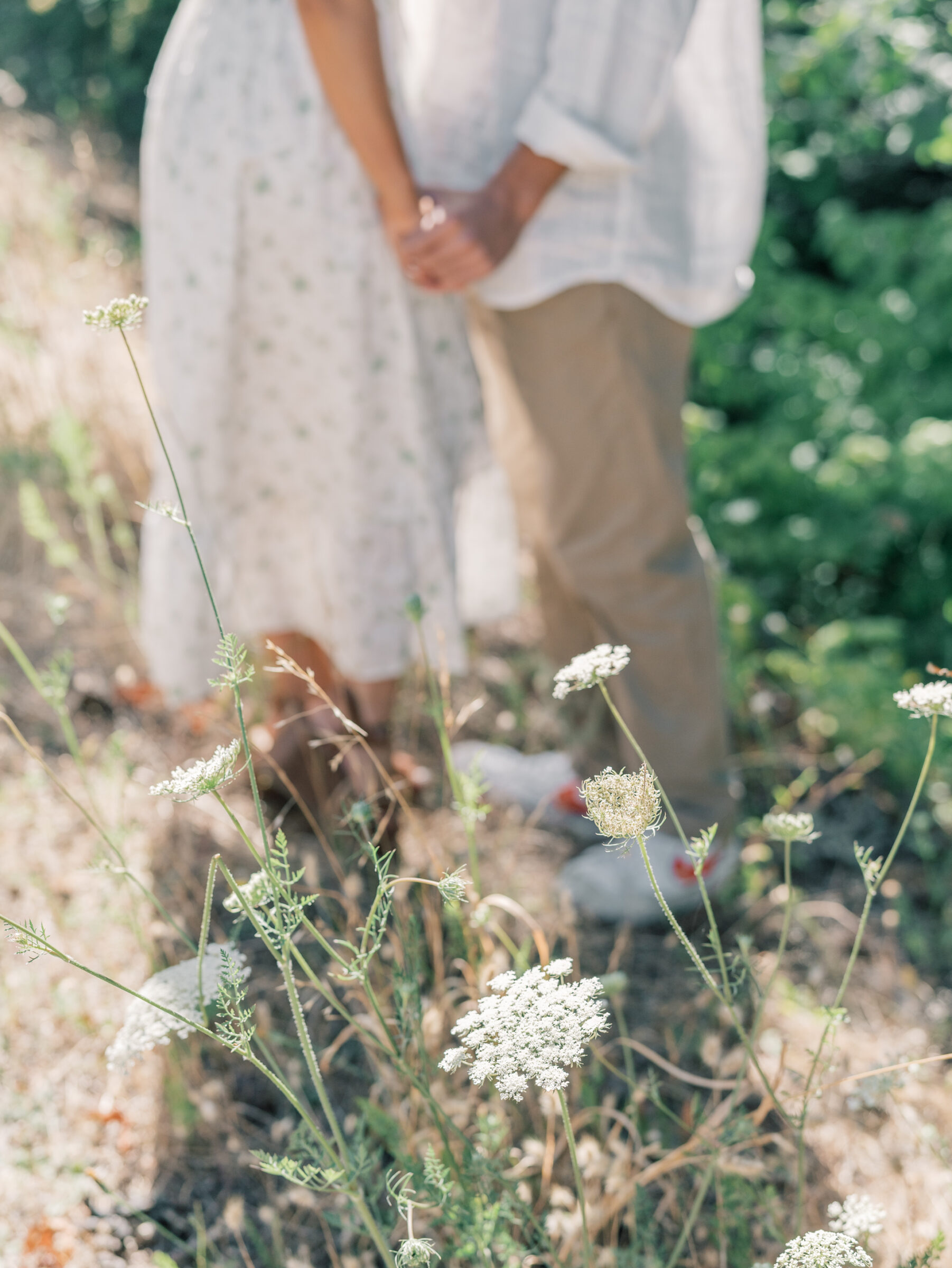 Wildflowers in focus with an engaged couple holding hands softly blurred in the background during their engagement session in Oregon.