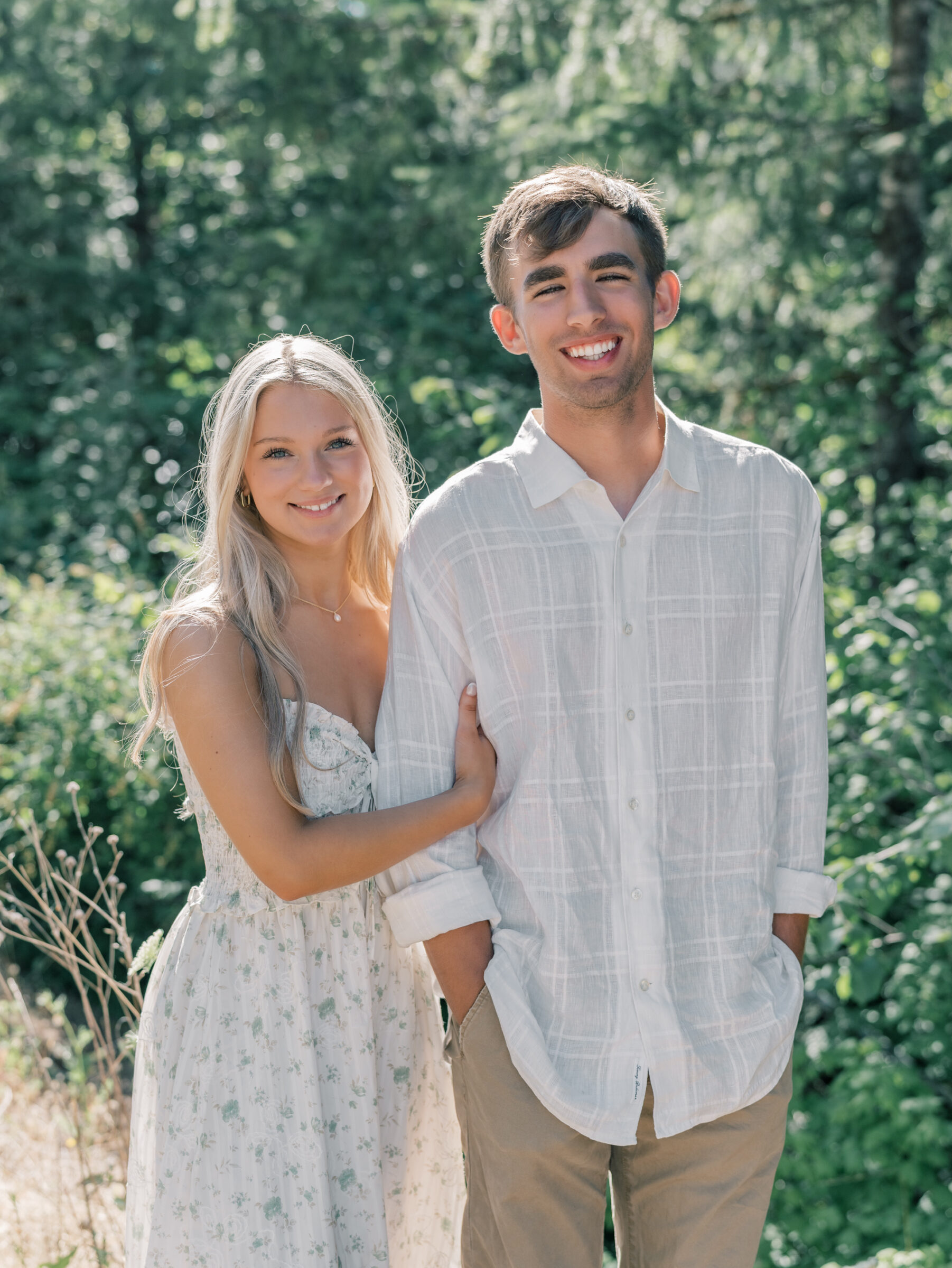 Smiling couple posing together during their Umpqua River engagement session in Oregon, with sunlit greenery and forest in the background.
