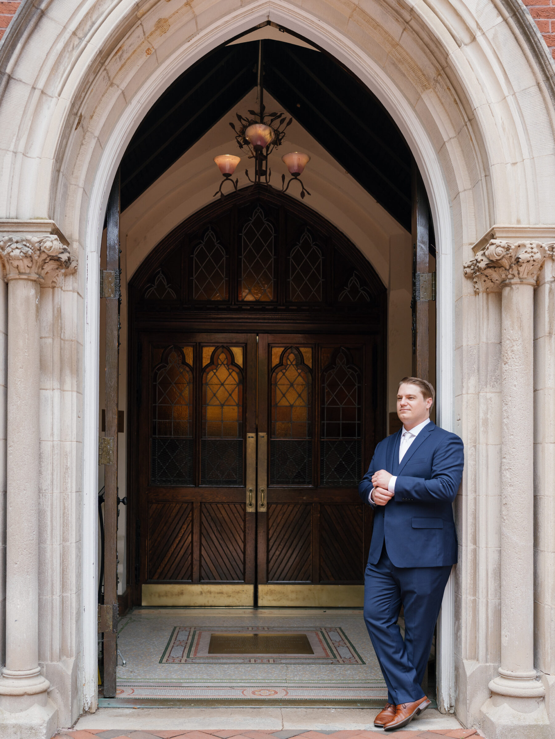 Groom in a navy suit standing outside the arched stone entryway of Dahlgren Chapel at Georgetown University before a Catholic wedding ceremony in Washington DC.