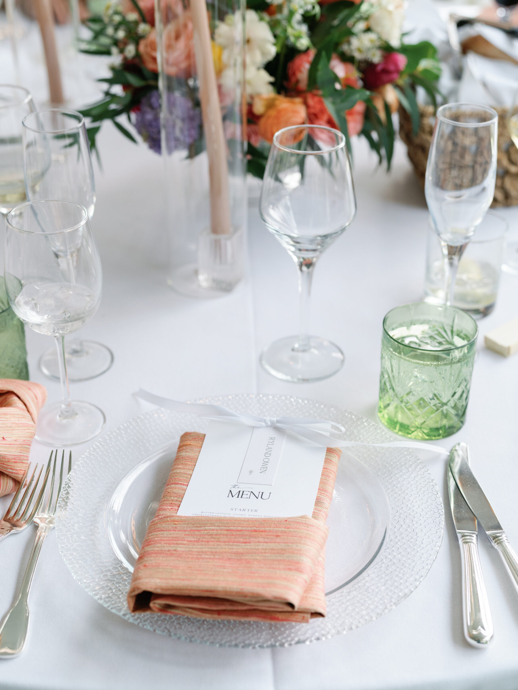 Elegant place setting with glass charger, coral napkin, and printed menu surrounded by floral centerpieces and glassware at a Woodrow Wilson House Wedding reception in Washington DC.