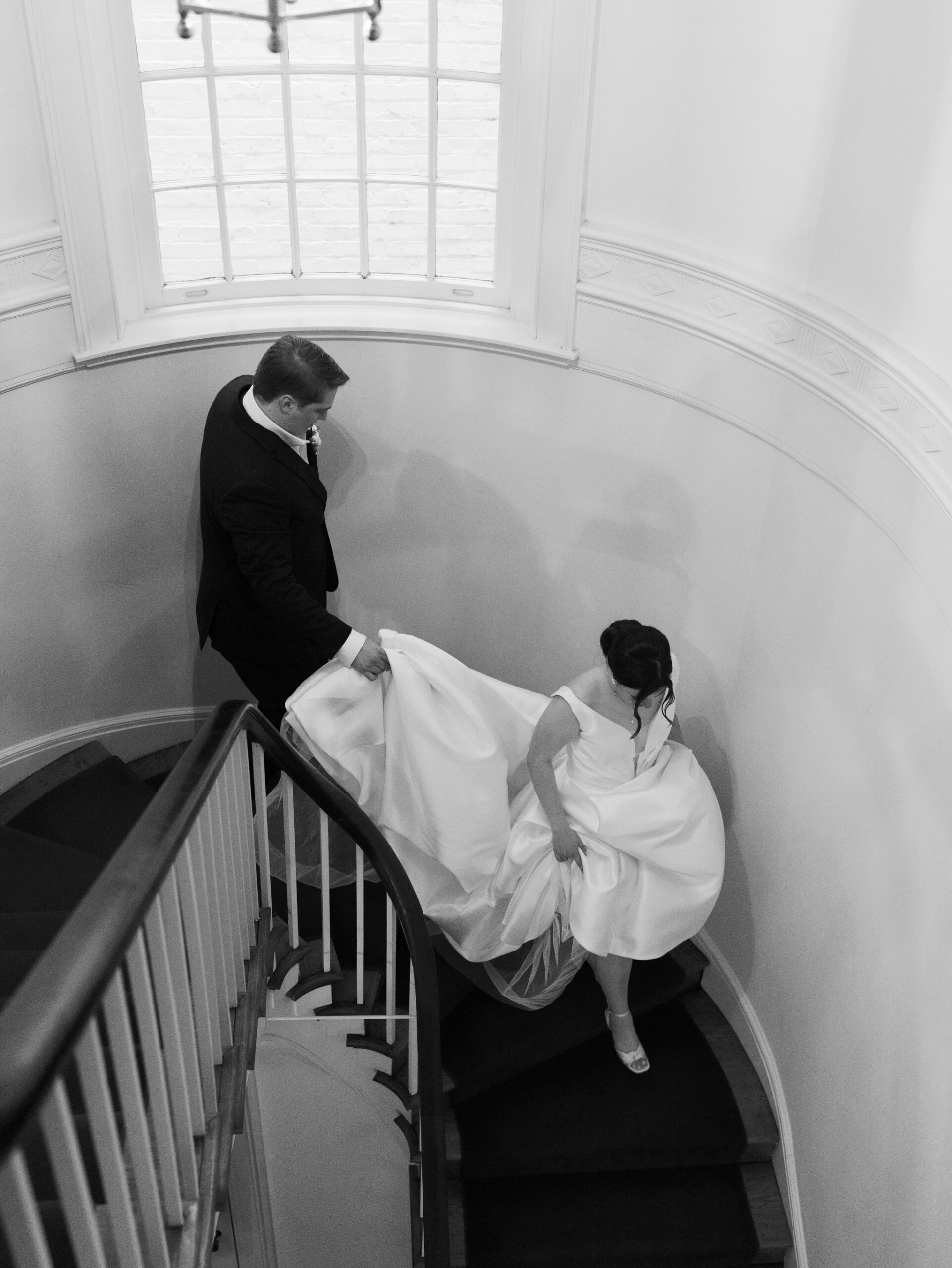 Bride and groom descend the curved staircase at the historic Woodrow Wilson House wedding venue in Washington DC, with the groom holding the bride’s train.