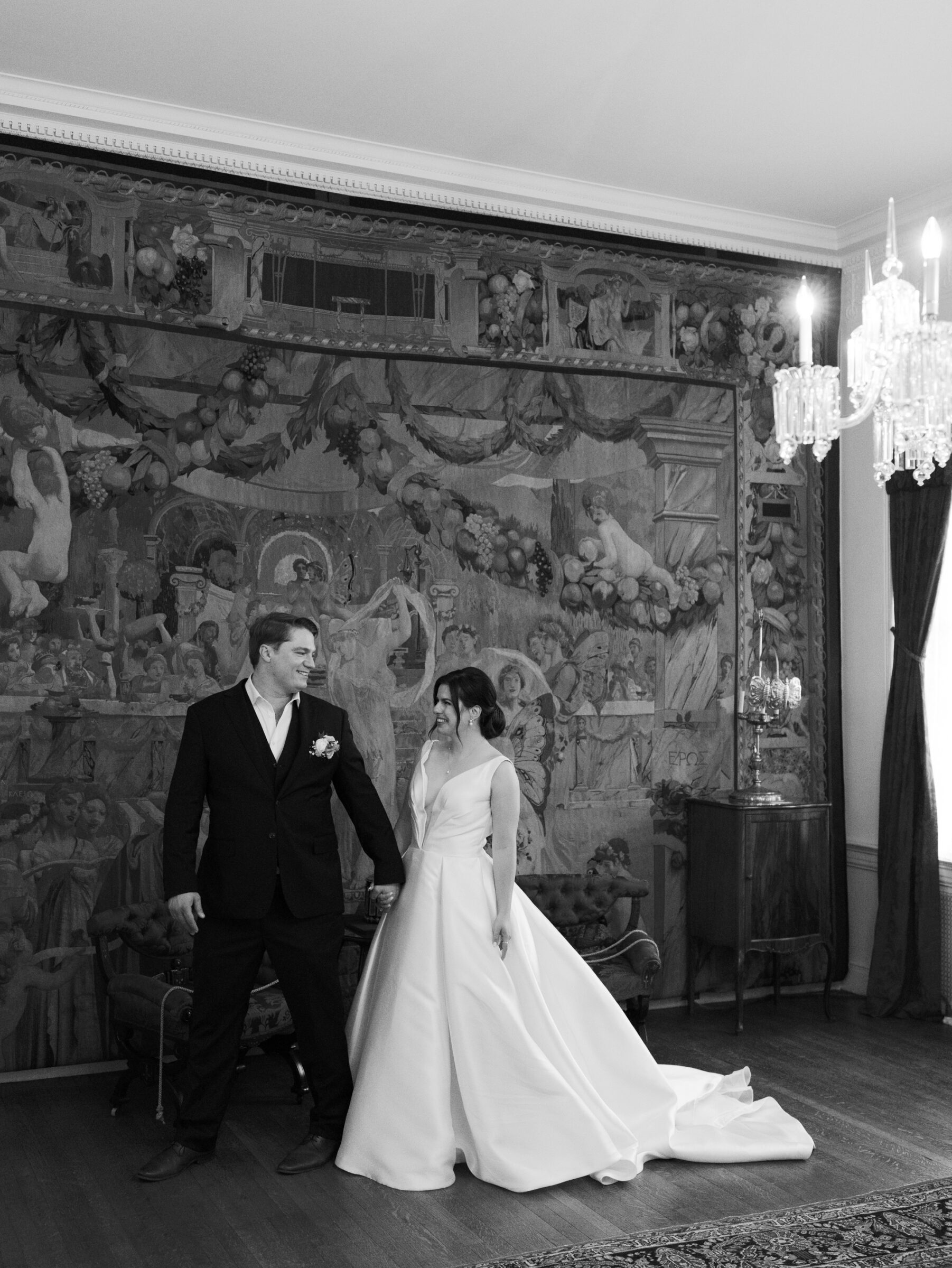 Bride and groom holding hands inside the historic Woodrow Wilson House Wedding venue in Washington DC, standing before a grand mural and chandelier.