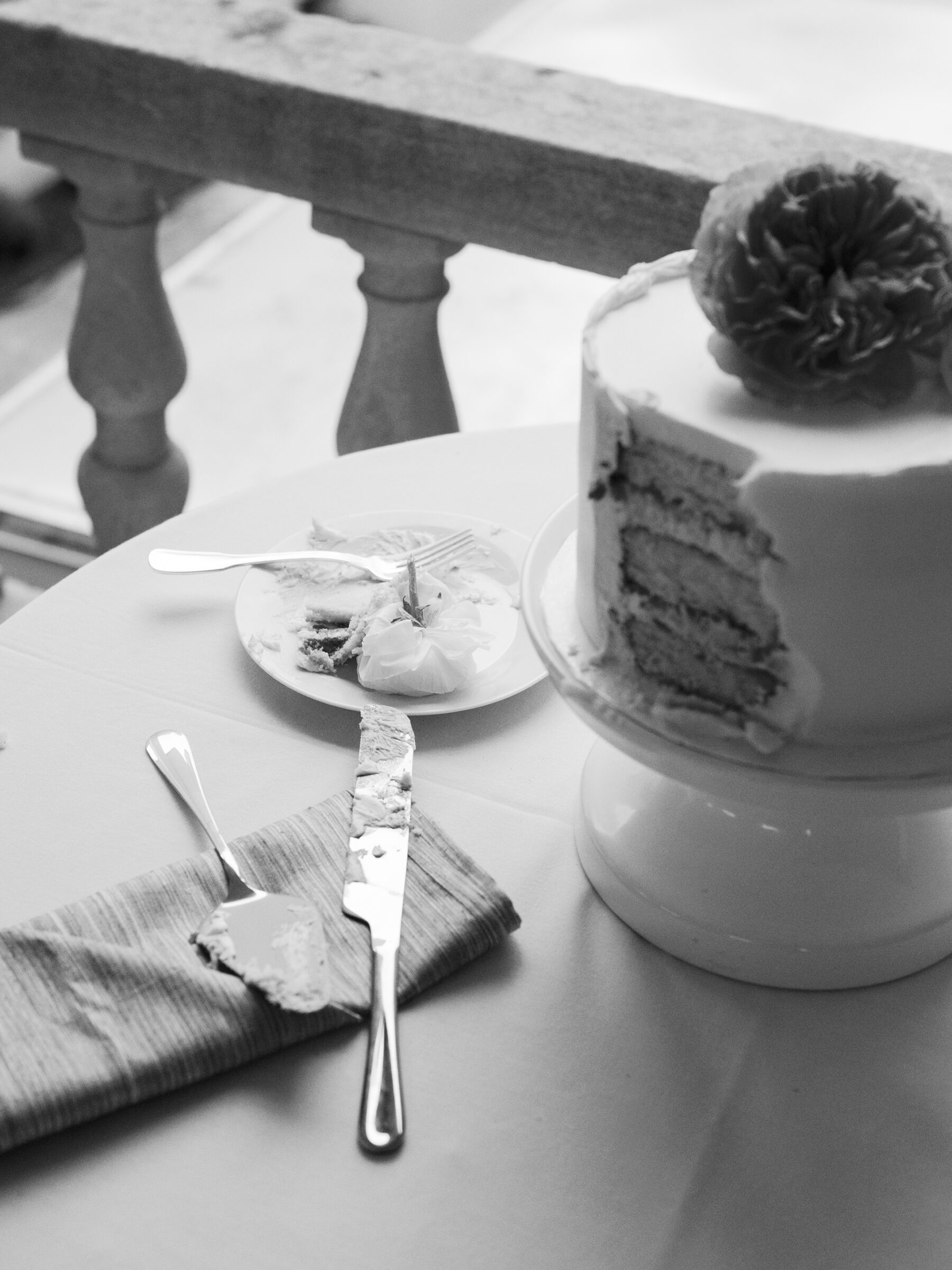 Black and white photo of a wedding cake with a slice cut out, displayed on a white pedestal with floral decoration at Woodrow Wilson House wedding in Washington DC.