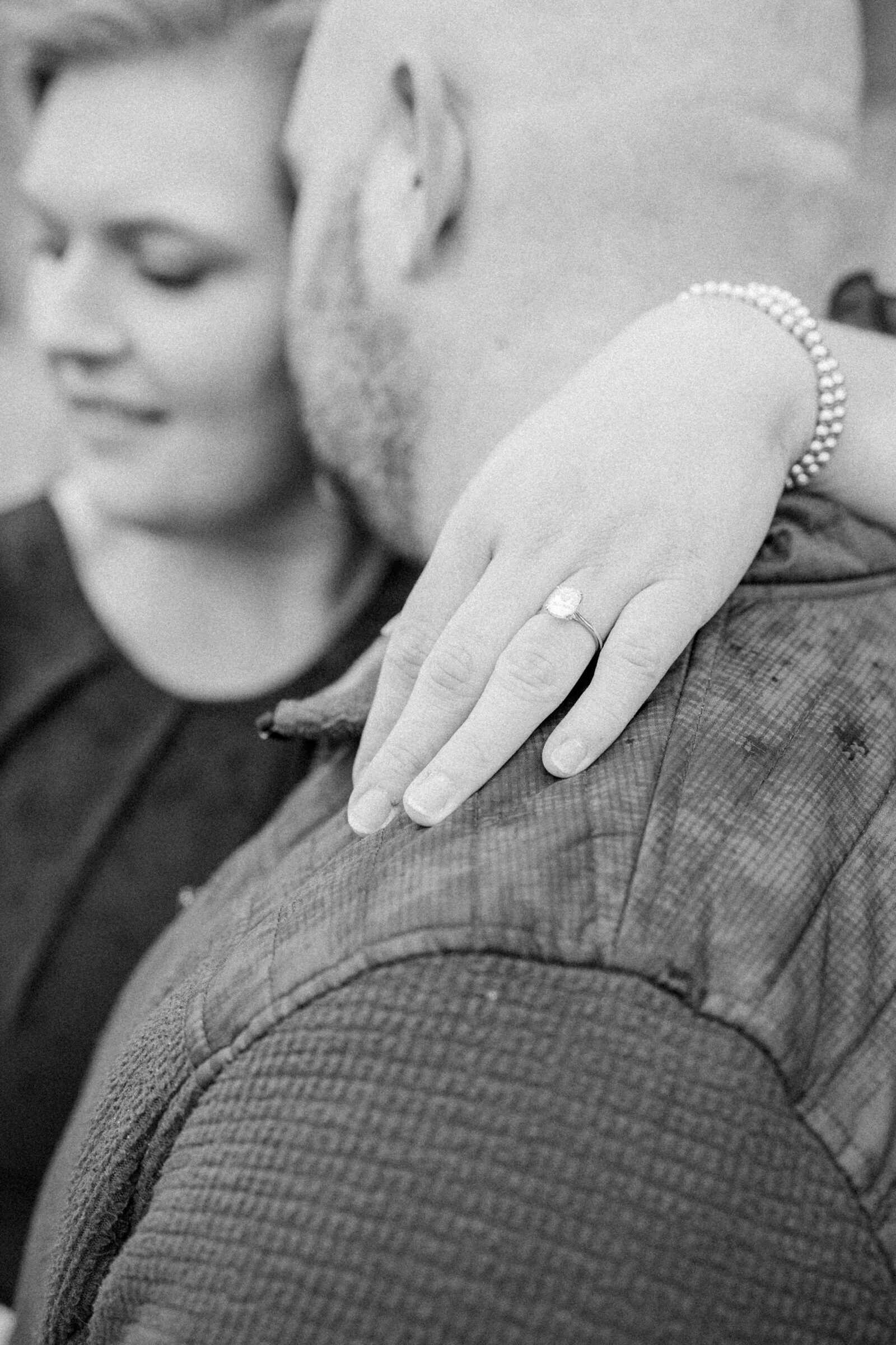 Black and white portrait of a couple, highlighting the bride’s engagement ring as she rests her hand on her fiancé’s shoulder.