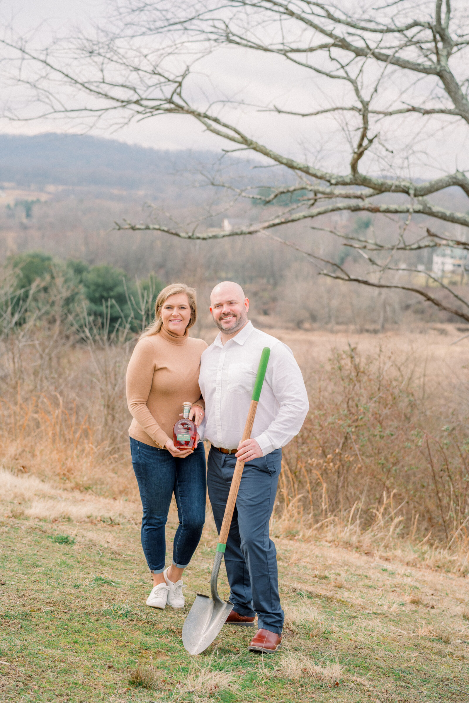 Couple holding a bourbon bottle and shovel during their bourbon burying ceremony at Blue Valley Vineyard in Delaplane VA with mountain views in the background.