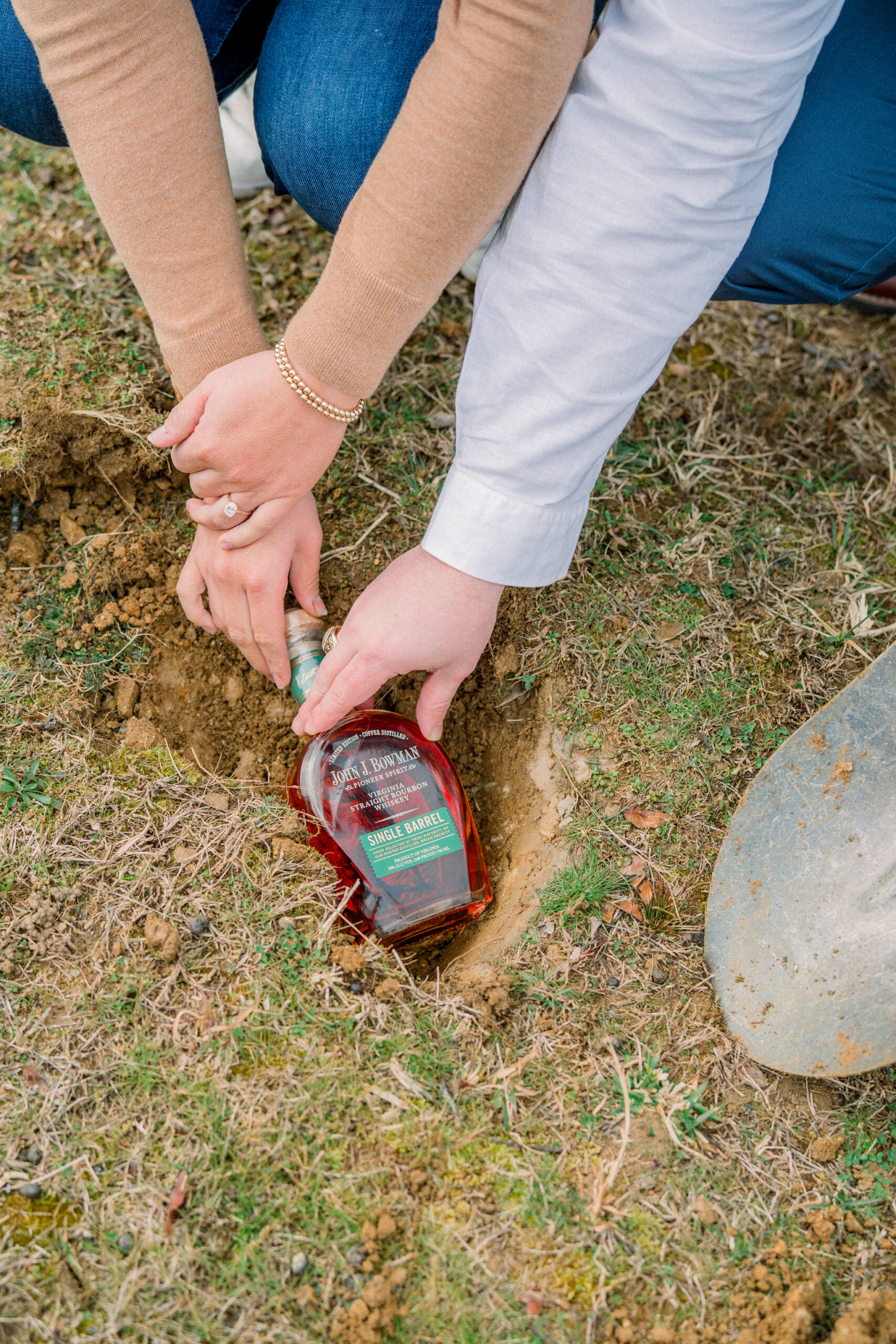 Close-up of a couple burying a bottle of bourbon during their bourbon burying ceremony at Blue Valley Vineyard in Delaplane VA, with hands resting on the bottle in the ground.