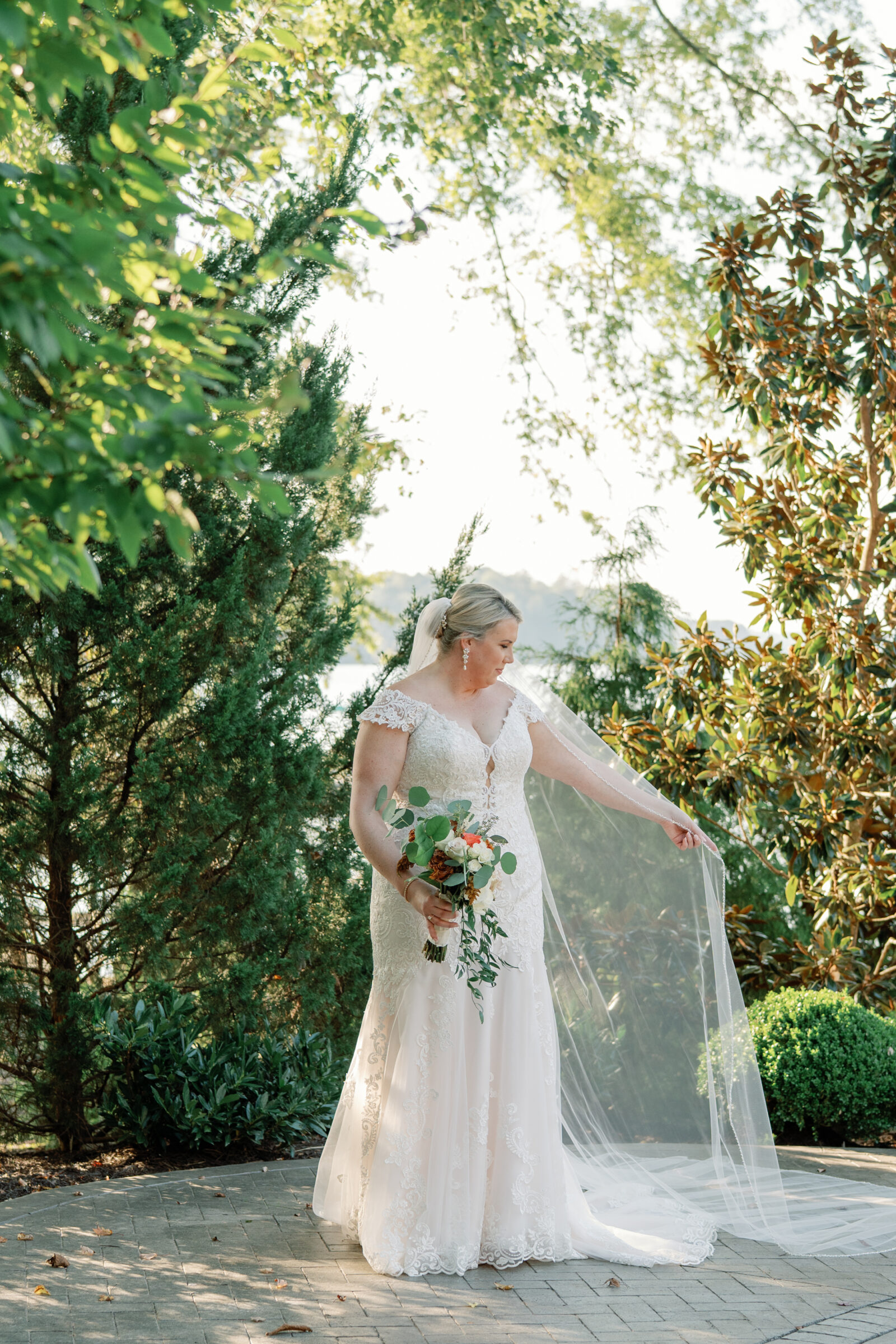 Bride in lace wedding gown holding a bouquet and lifting her veil during portraits at The Boathouse at Sunday Park wedding in Richmond VA.