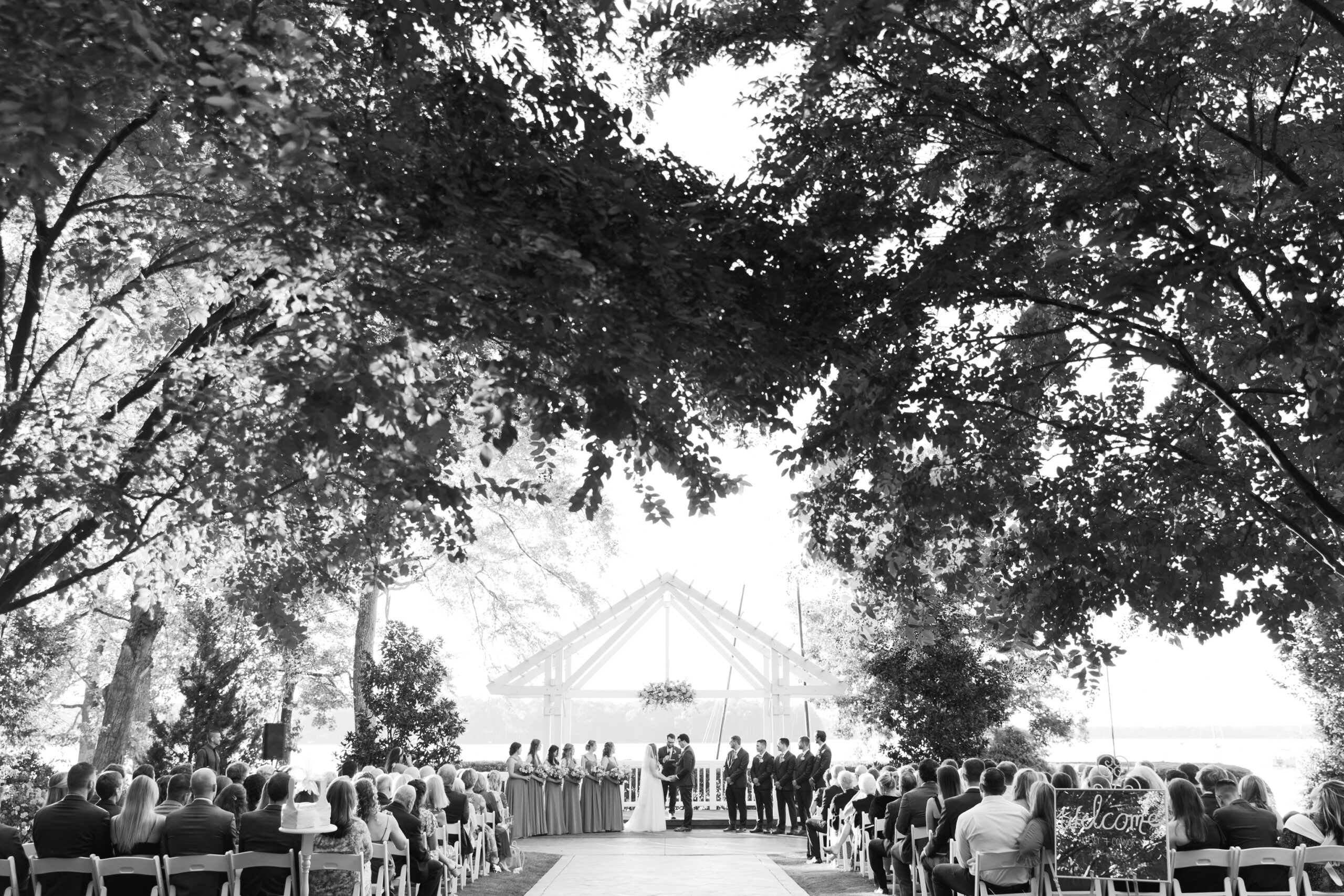 Black and white photo of a waterfront wedding ceremony at The Boathouse at Sunday Park in Richmond VA, with guests seated under trees and the couple exchanging vows beneath a wooden arbor.