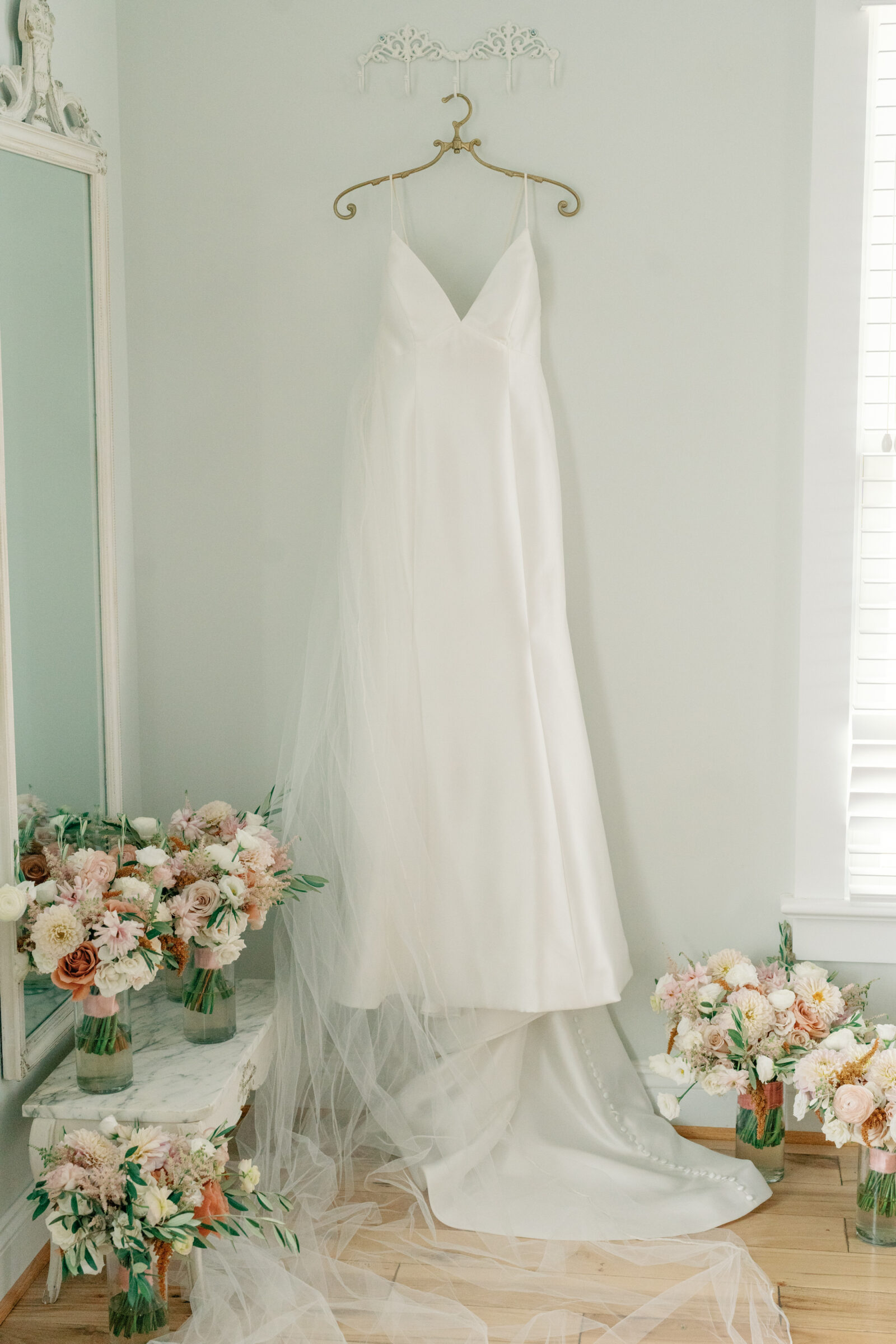 Elegant silk wedding gown with a flowing veil hanging in the bridal suite at Burlington Weddings & Events in Charles City VA, surrounded by pastel floral bouquets.