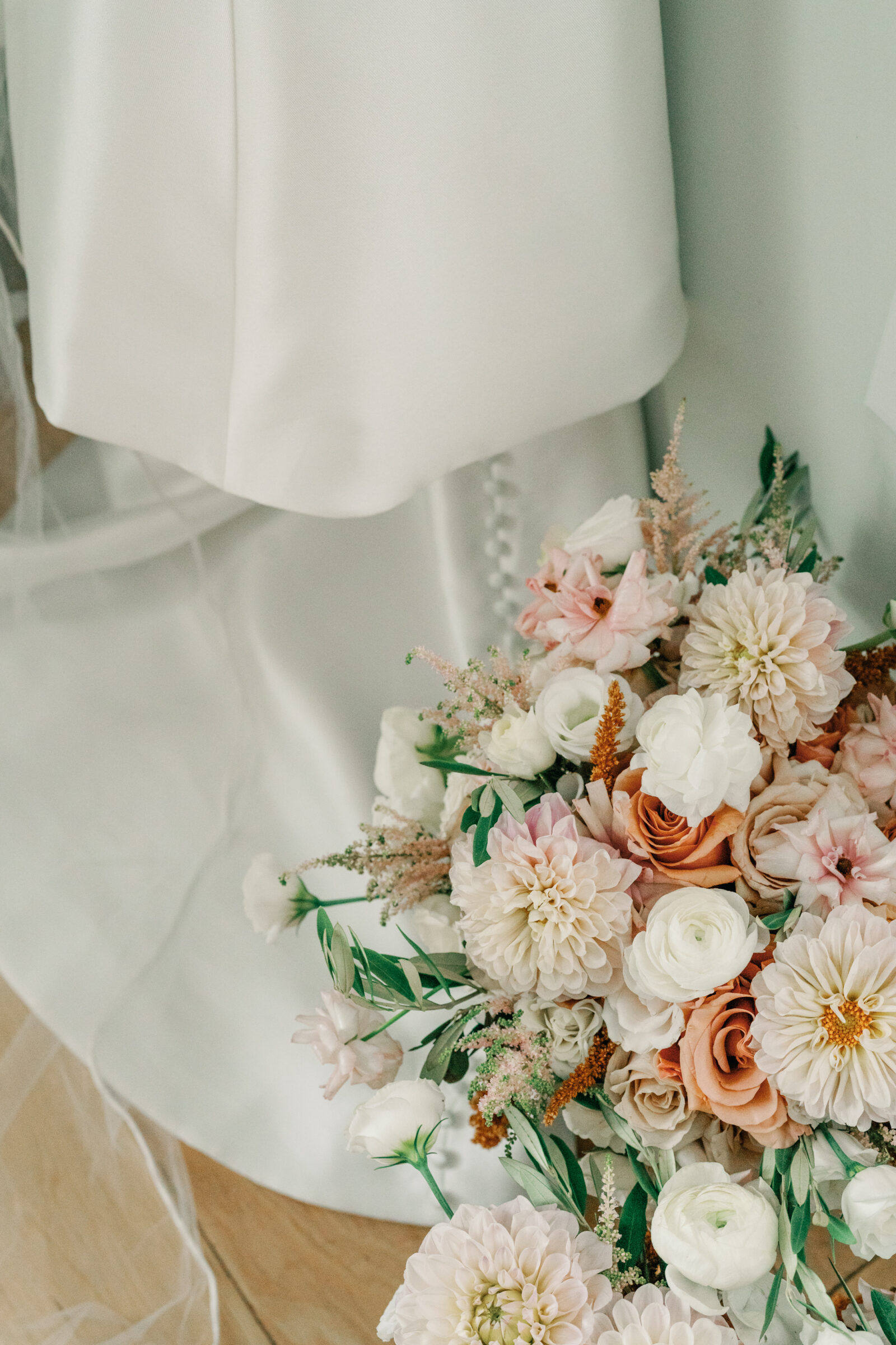 Close-up of pastel bridal bouquet with dahlias, roses, and ranunculus placed beneath a silk wedding dress.